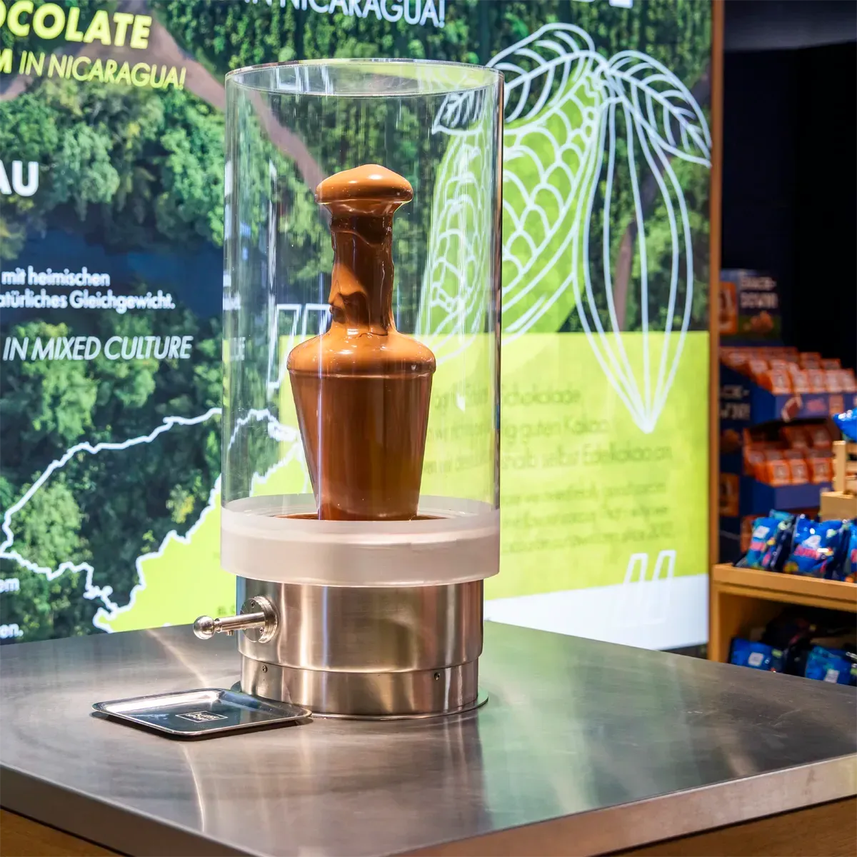 Ein Schokoladenbrunnen in einer Glasvitrine auf einer Metalltheke, im Hintergrund ein Informationsplakat.