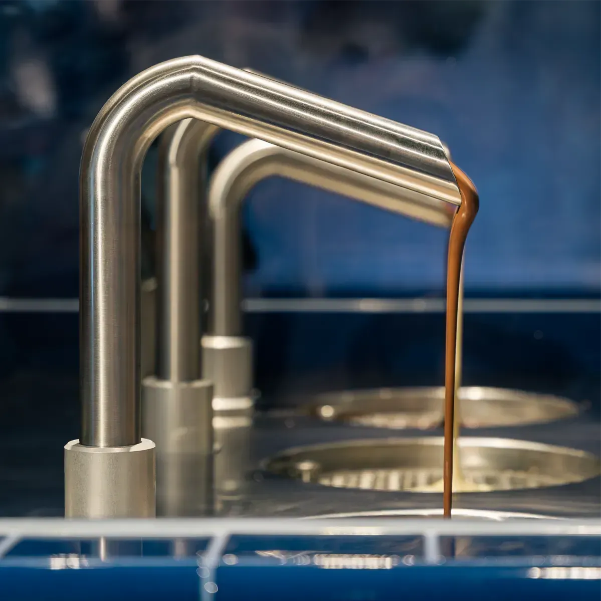 Ein metallener Ausguss ergießt einen Strahl flüssiger Schokolade in einen Abflussrost vor einem dunkelblauen, gefliesten Hintergrund.