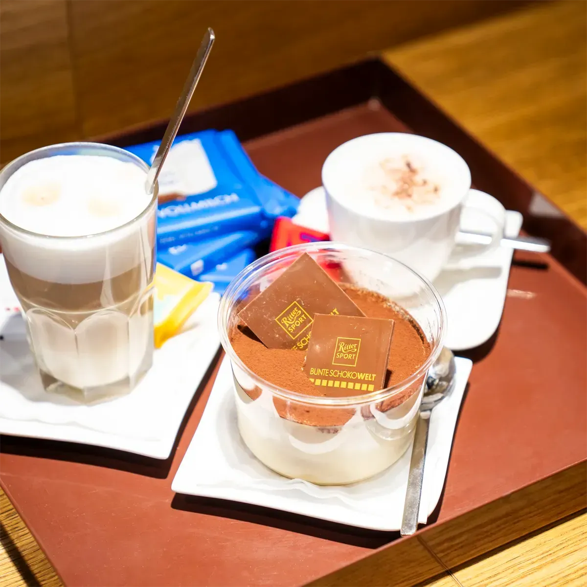 Auf einem dunklen Tablett werden zwei Kaffeegetränke und ein Tiramisu-Dessert mit Schokoladenquadraten serviert.