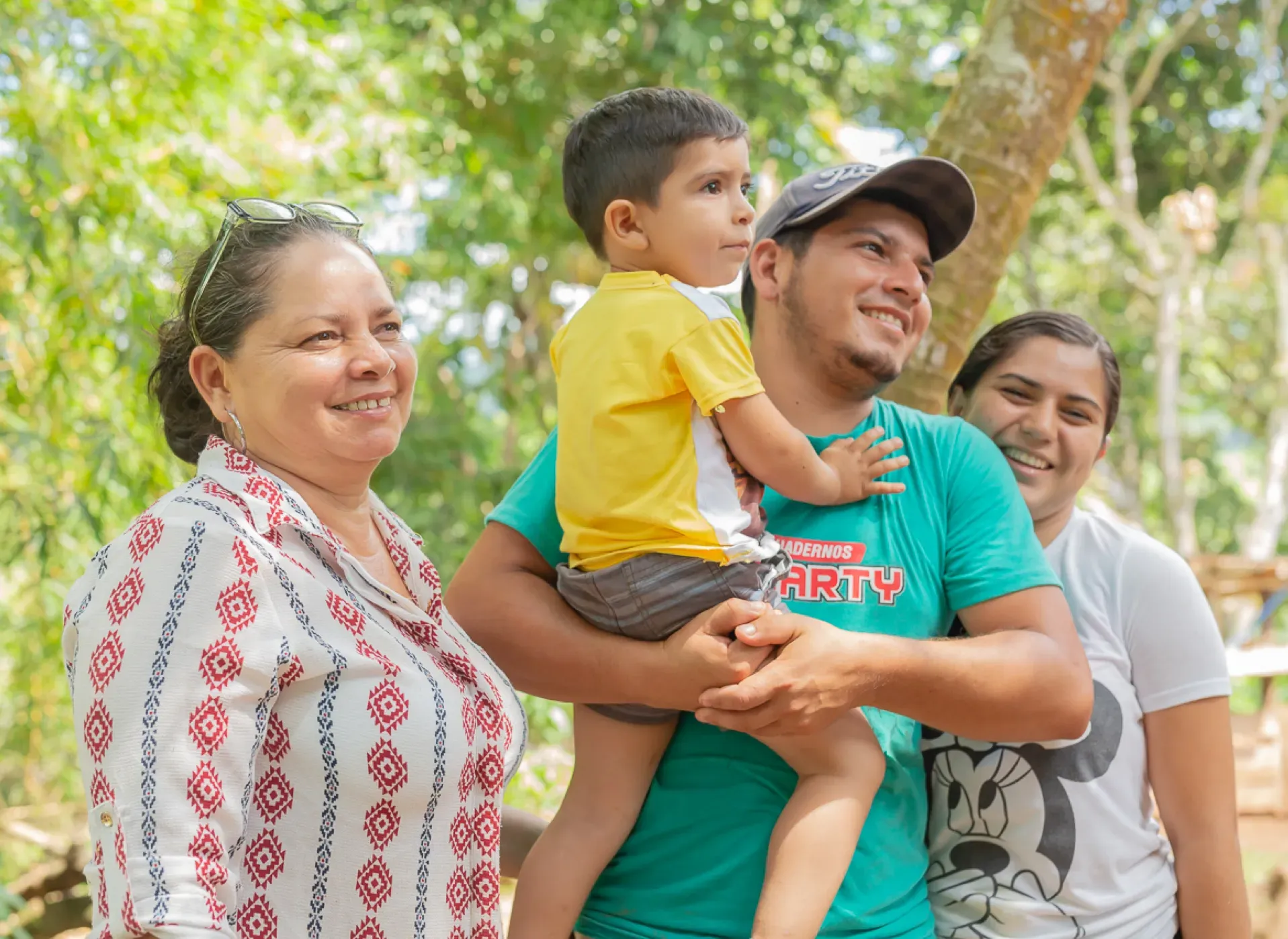 Family smiling outdoors: A man holding a child, two women, trees in the background.