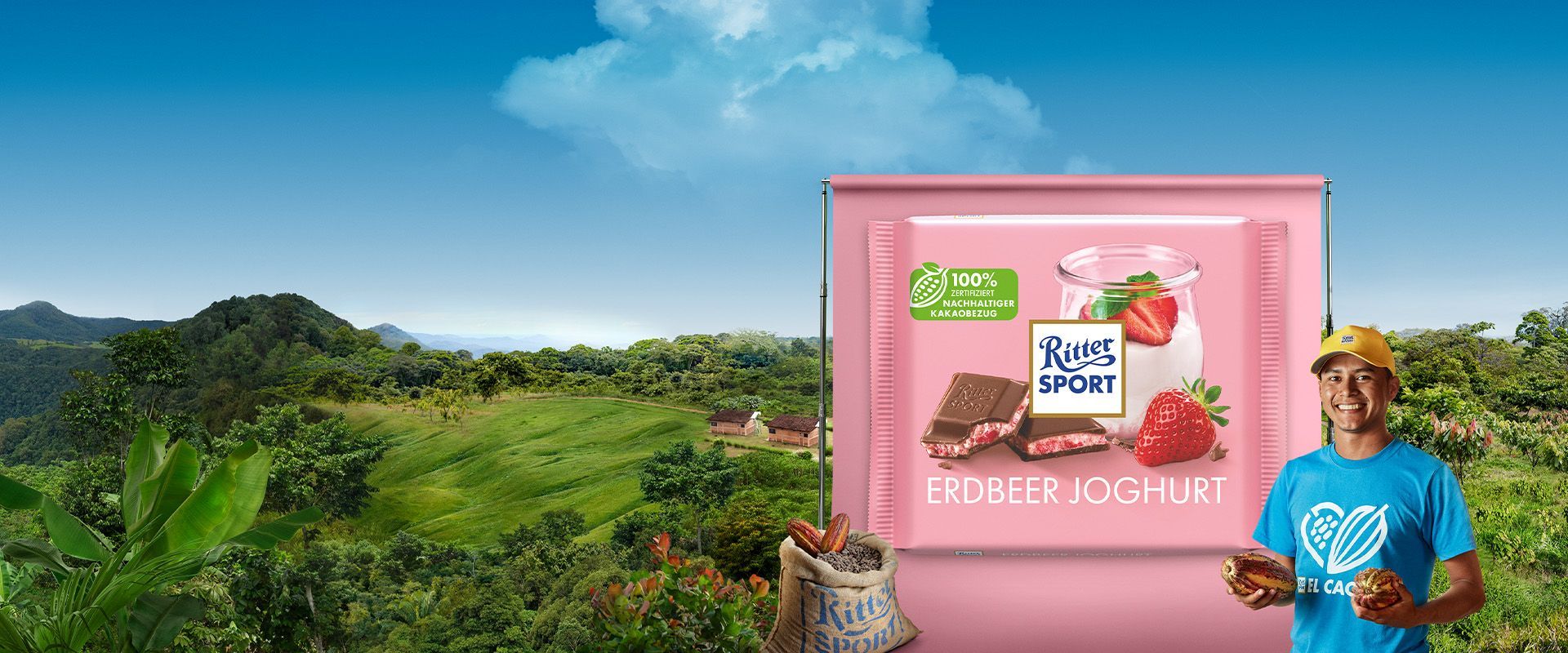 Ein Mann im blauen Hemd hält eineKakaobohne. Vor einer grünen Landschaft ist eine Schokolade mit Erdbeer-Joghurt ausgestellt.
