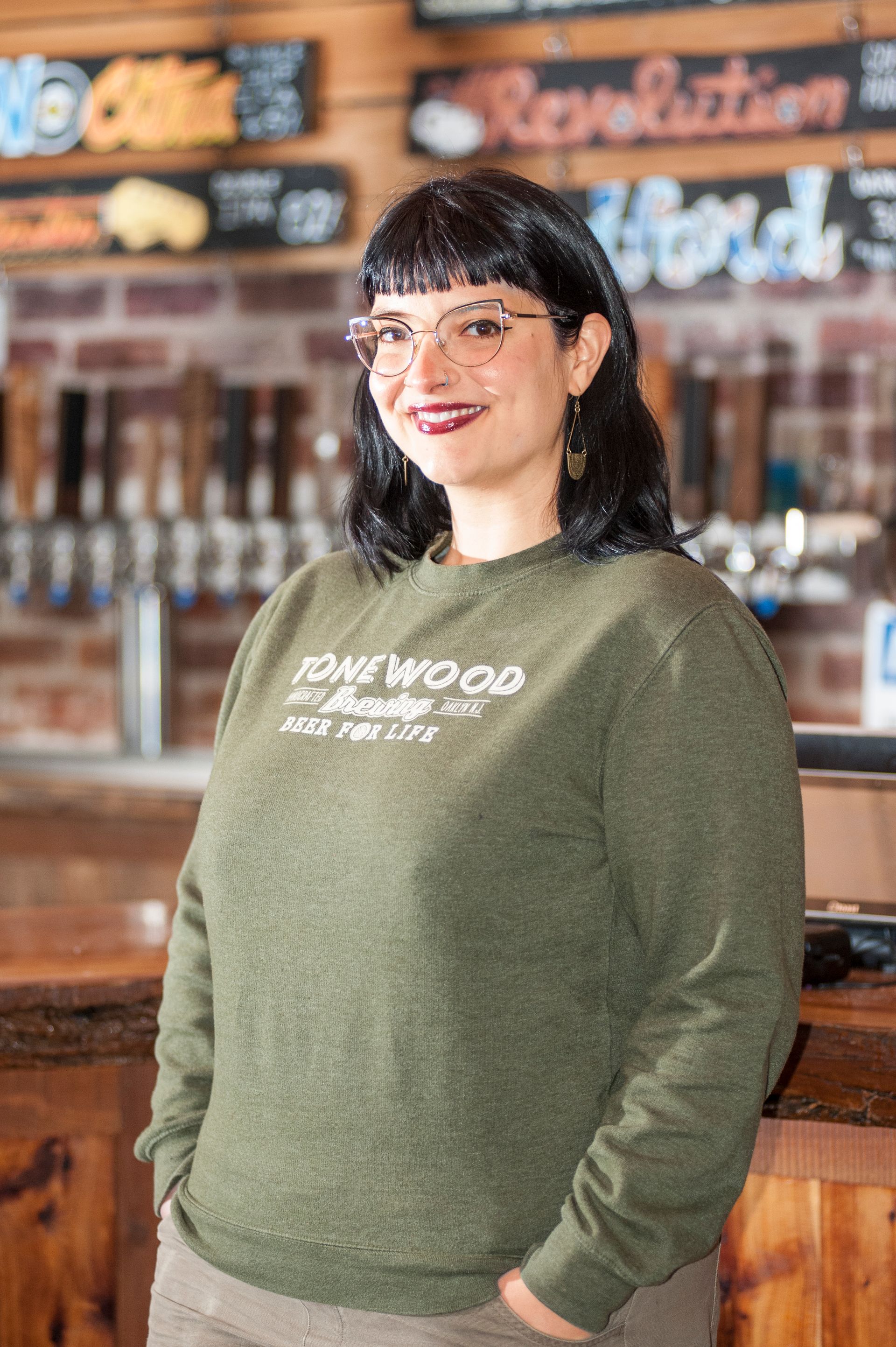 Woman with glasses wearing a green sweatshirt, leaning on a bar in a brewery.
