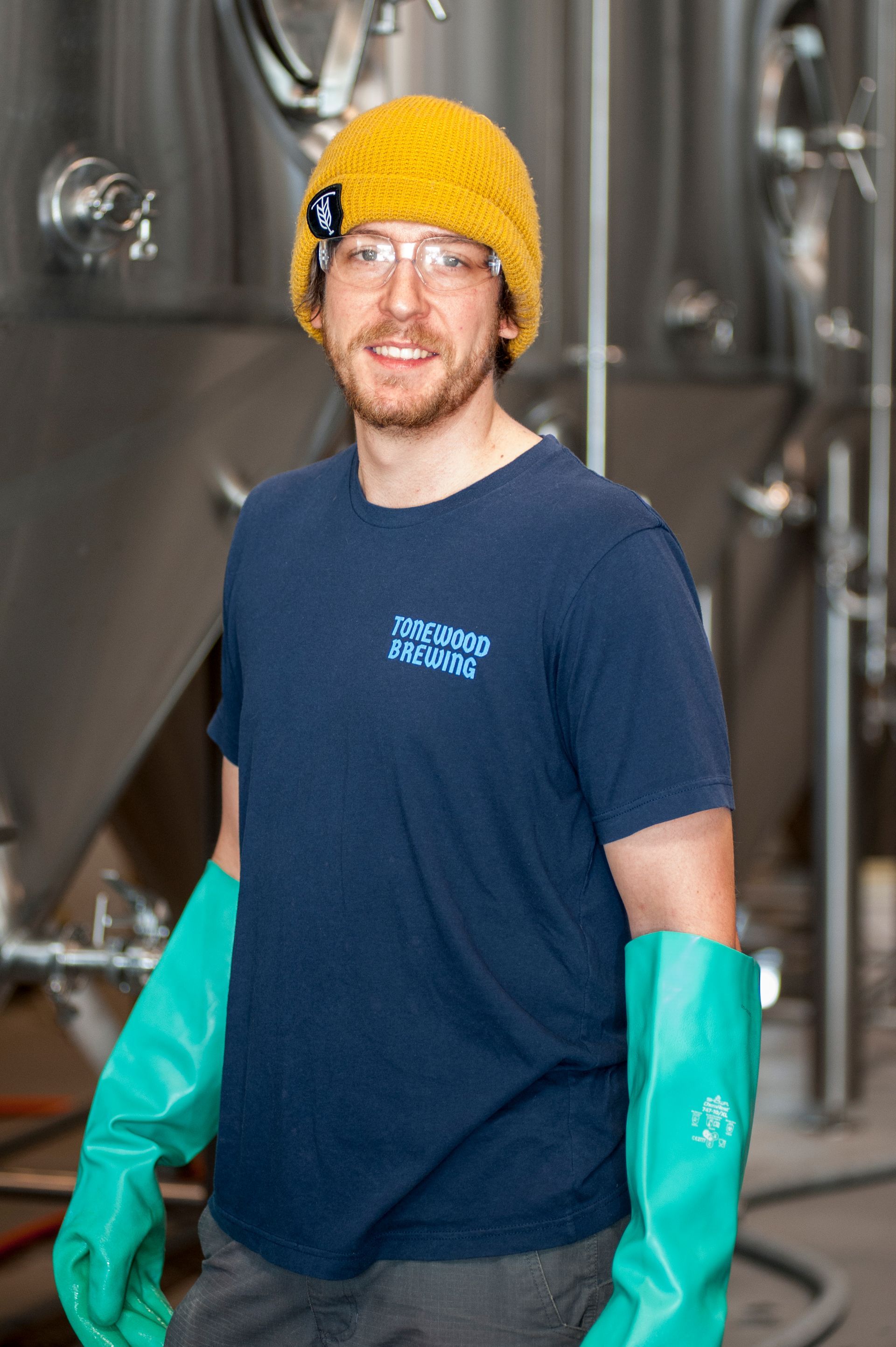Man in yellow hat and blue shirt smiles, wearing green gloves in a brewery.