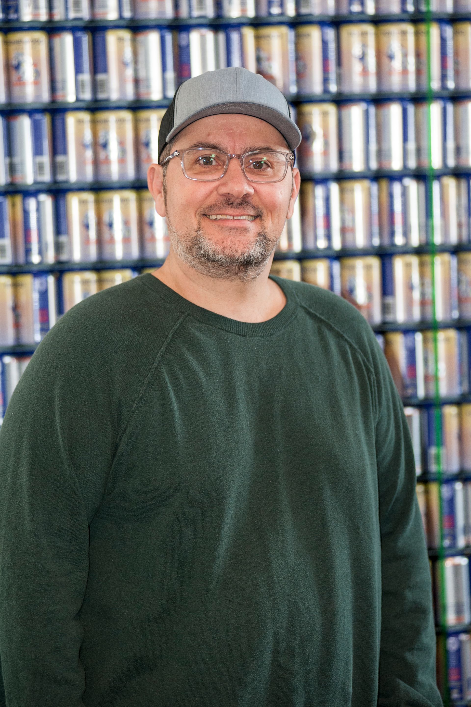 Man in green sweater and hat smiles, standing in front of a wall of Red Bull cans.