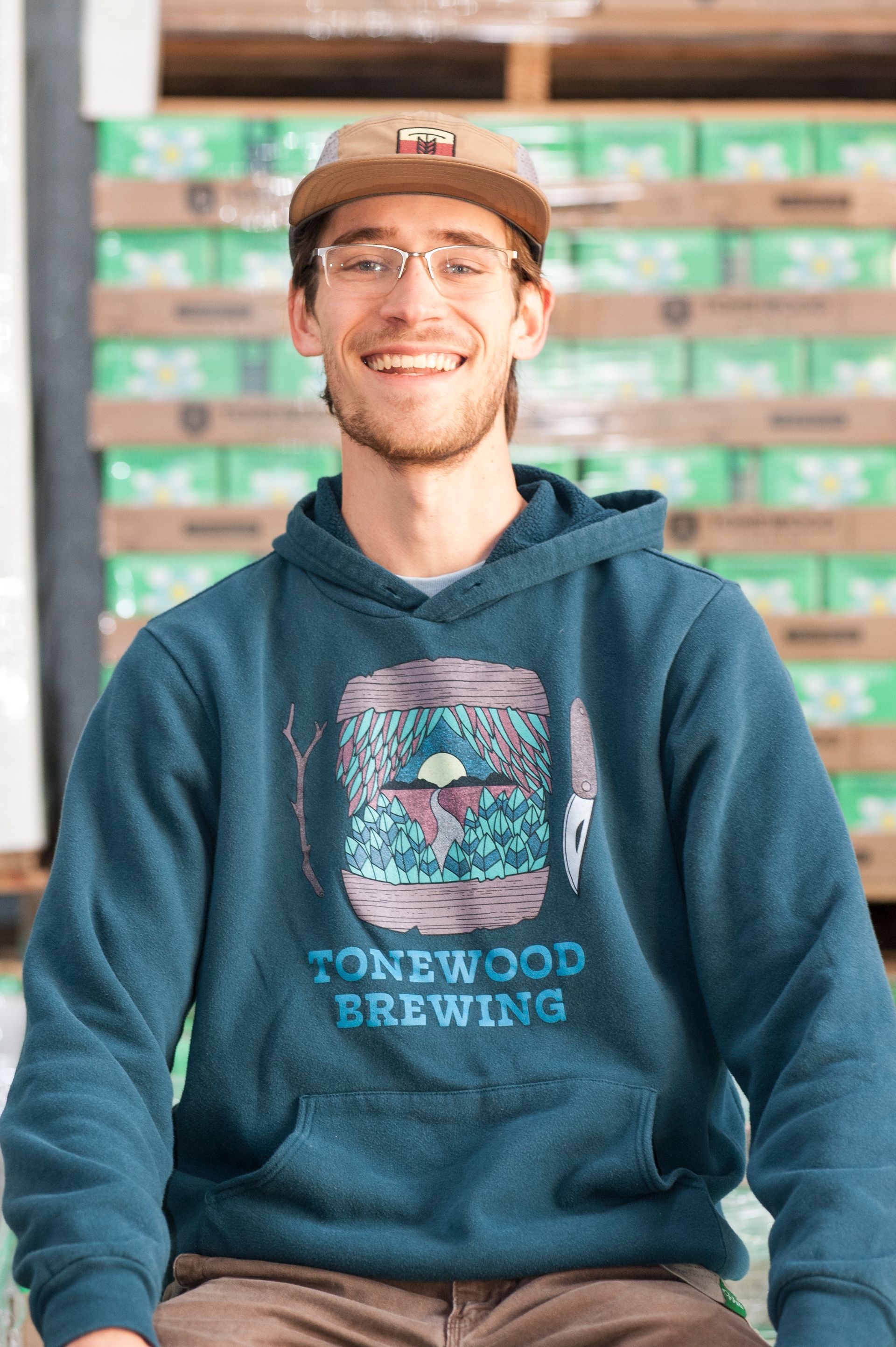 Man wearing Tonewood Brewing hoodie smiles, posing near beer cases.