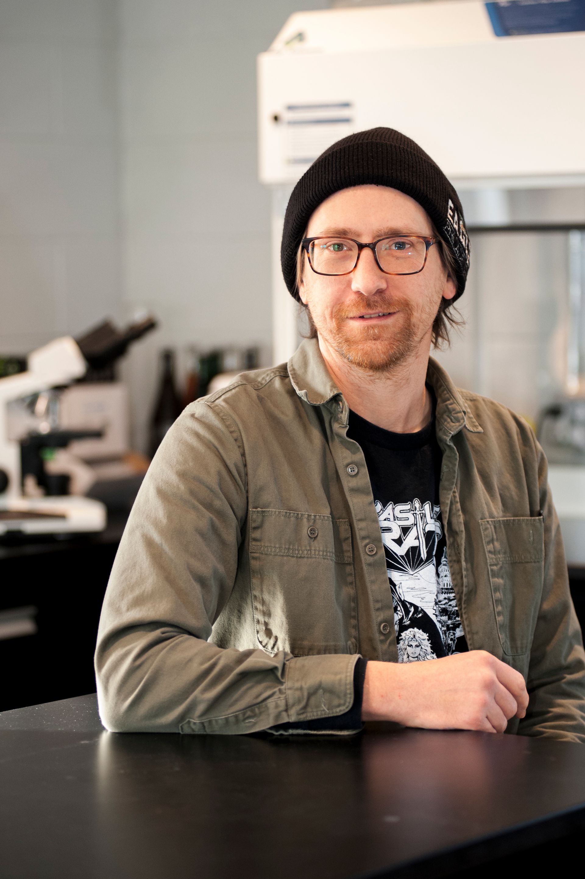 Man in glasses and black beanie in a lab setting, leaning on a table, smiling.