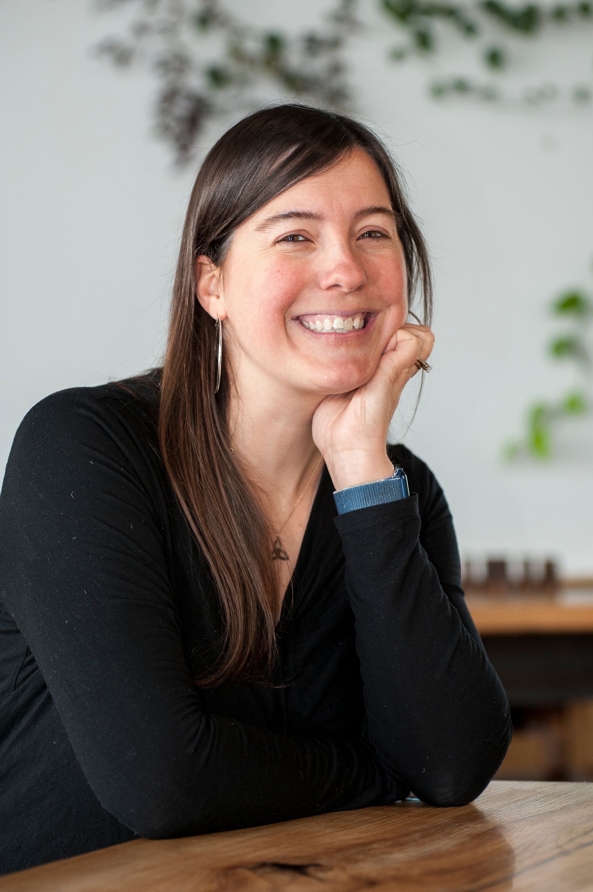 Woman smiling, resting chin on hand at a wooden table. Wearing a black sweater, indoor setting.