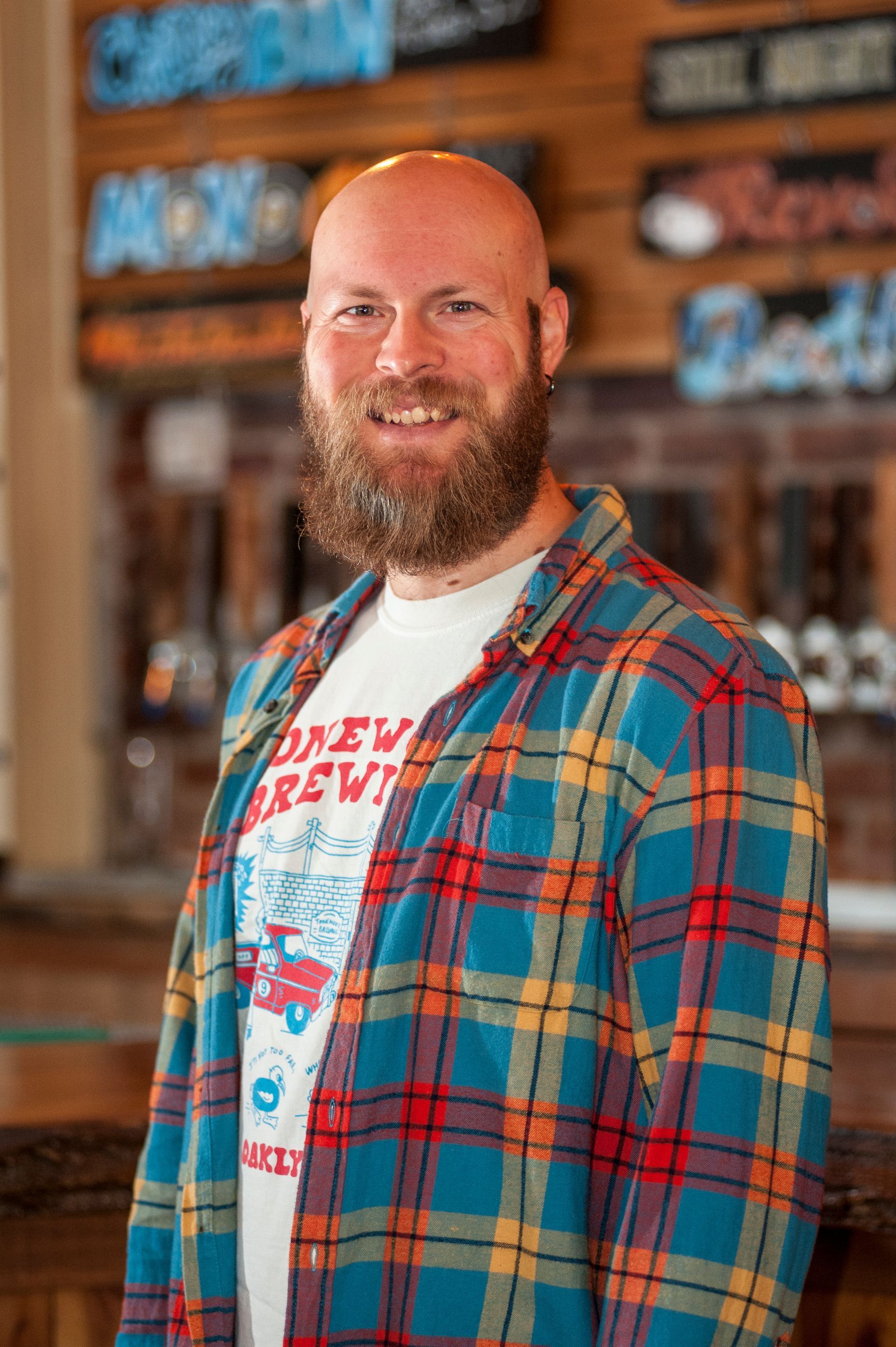 Man with a bald head and beard wearing a flannel shirt and smiling in a brewery.