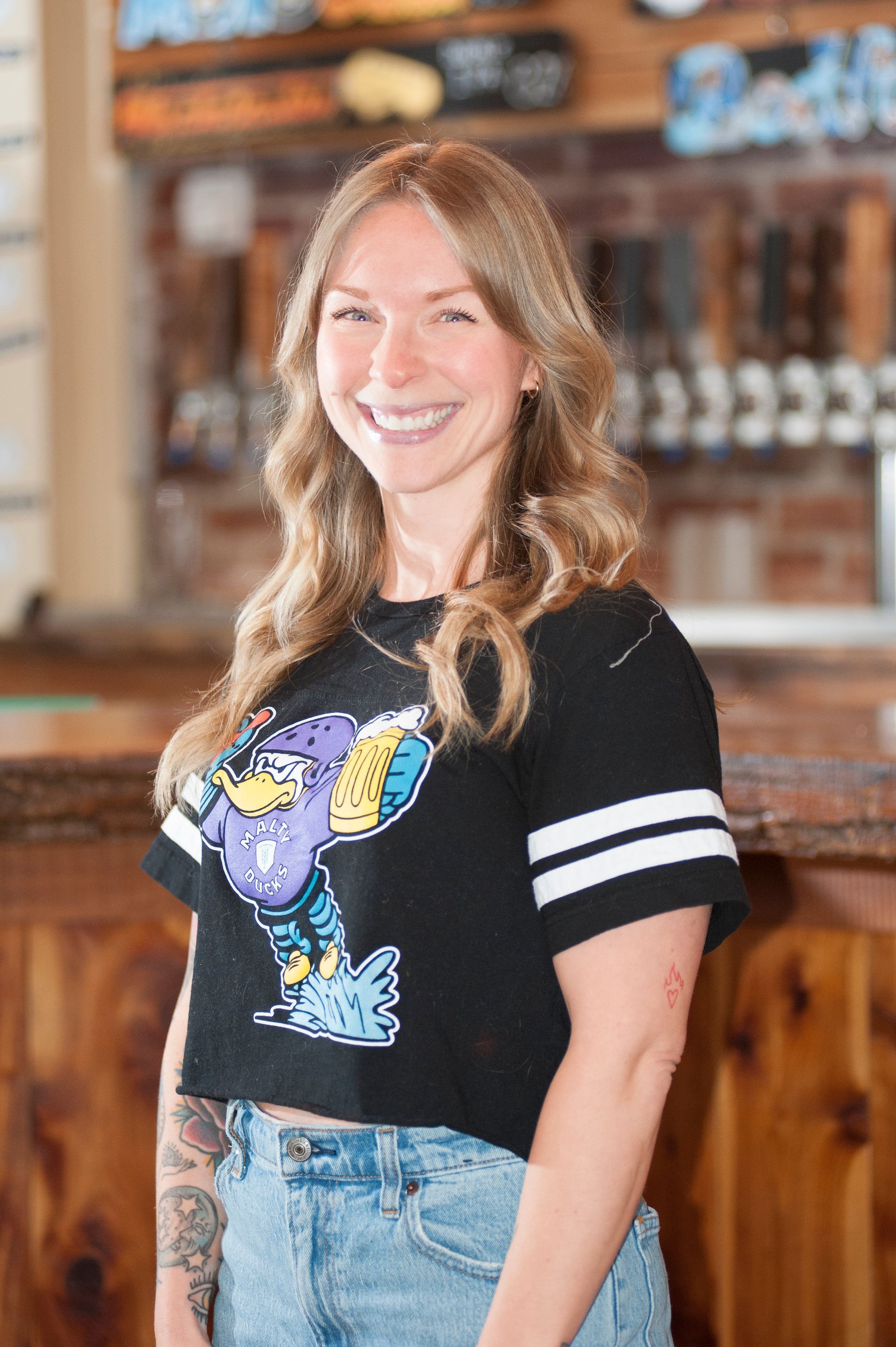 Woman smiling, wearing a black crop top with a purple cartoon, in a bar setting.