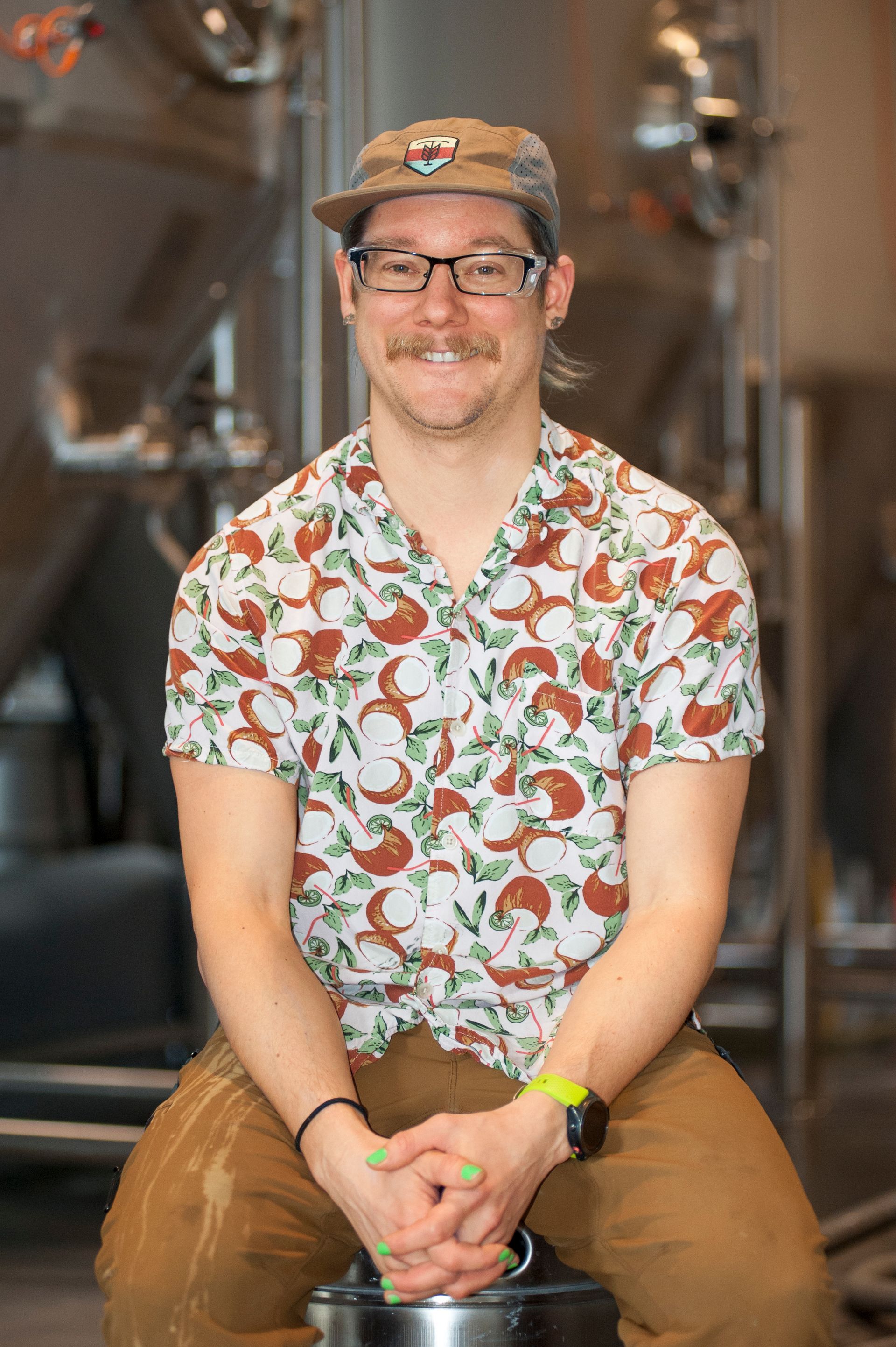 Man wearing glasses and hat, smiling, sitting. Wearing patterned shirt, in brewery setting.