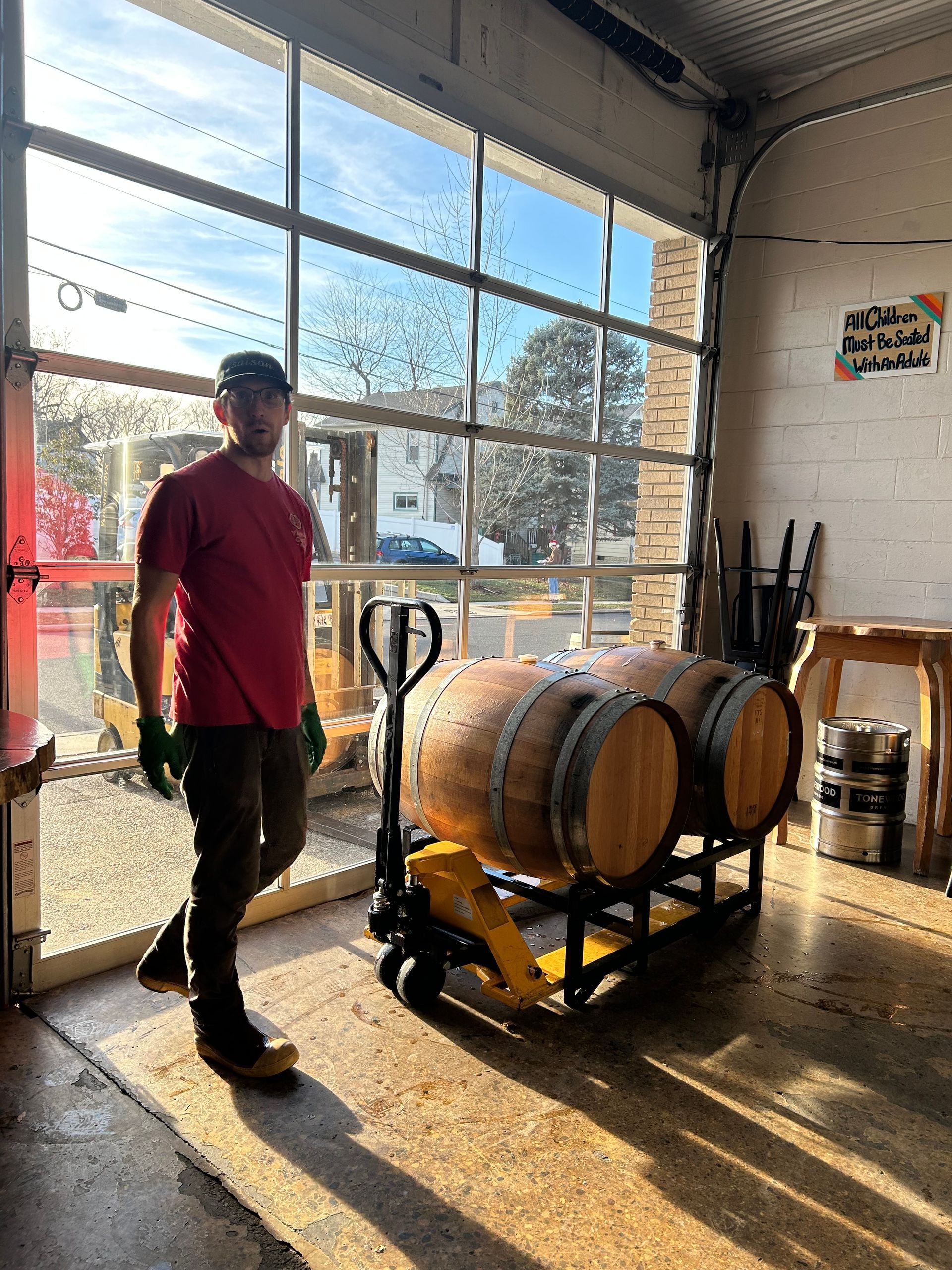 Man moving two wooden barrels on a pallet jack inside a building with a large glass door.