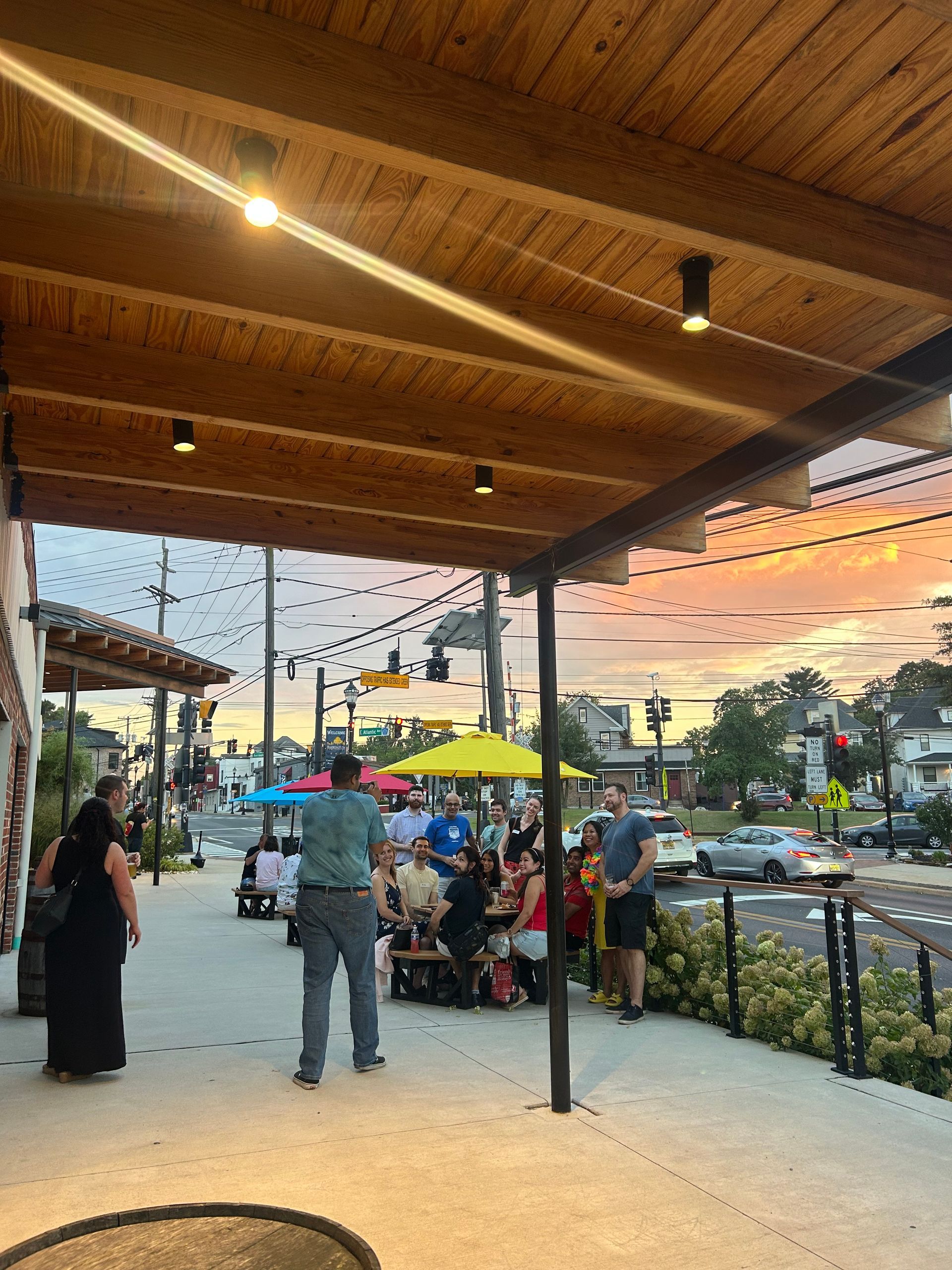 Outdoor patio with people dining under a wooden canopy. Strings of lights, street in the background. Sunset colors.