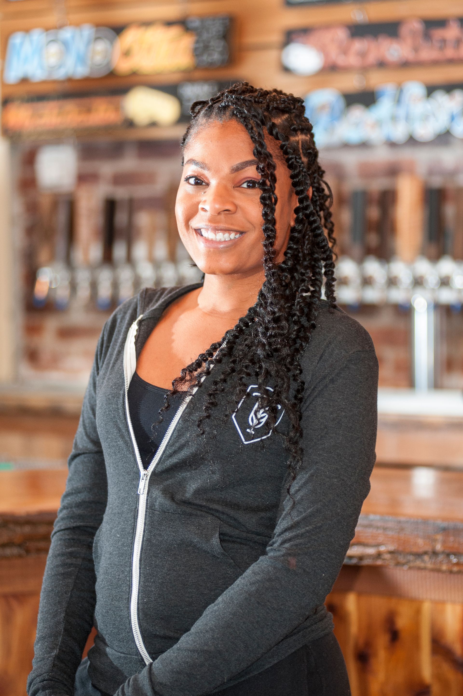Woman with long braids smiles, wearing a grey zip-up hoodie, standing in front of bar with taps and a brick wall.