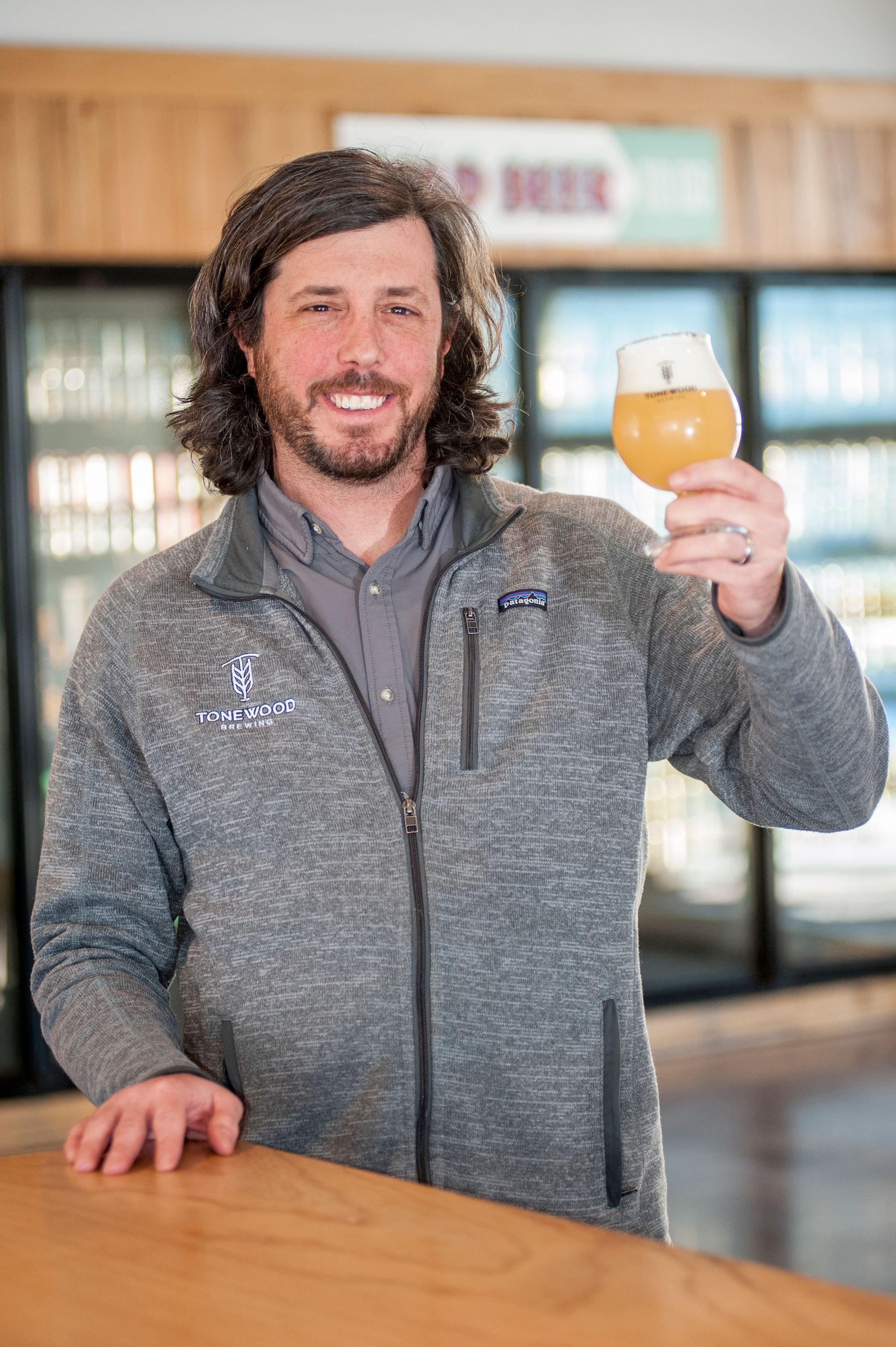 Man holding up a glass of beer, smiling. Wearing a gray zip-up sweater. Standing in a brewery setting.