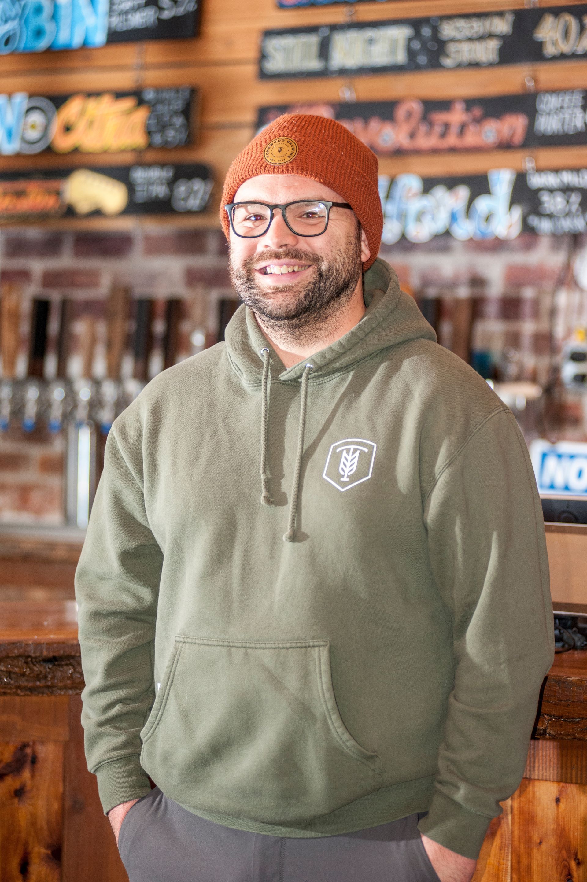 Man wearing a brown beanie and olive hoodie smiles inside a brewery, hands in pockets.