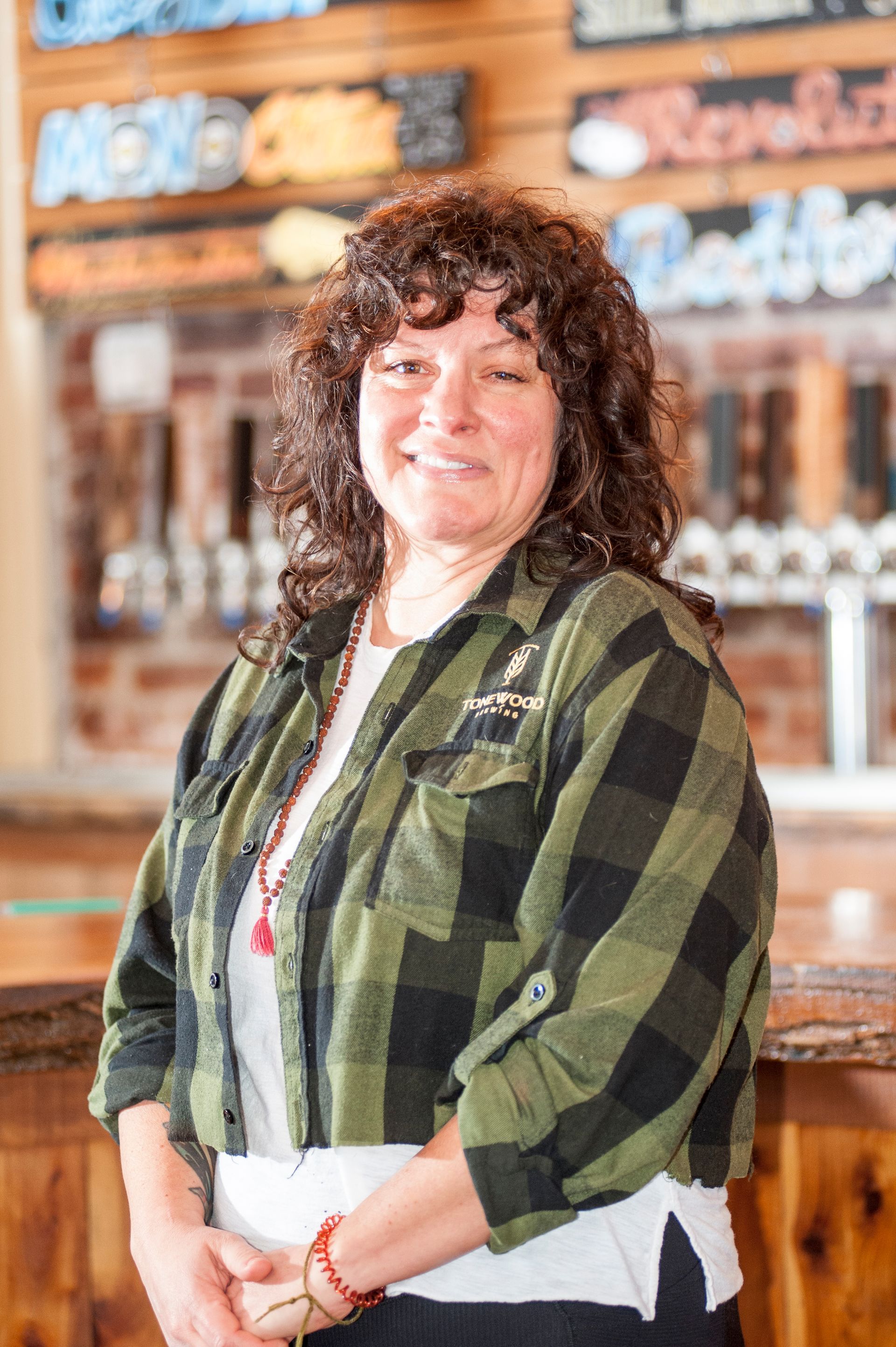 Woman in a green plaid shirt smiles in front of a bar, arms crossed.