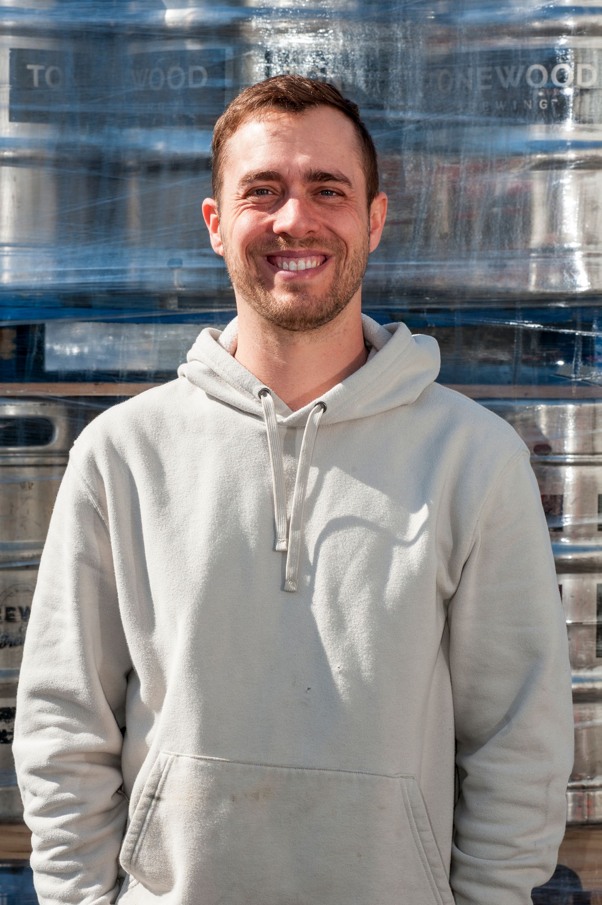 Man in a gray hoodie smiles in front of a stack of metal kegs, possibly at a brewery.