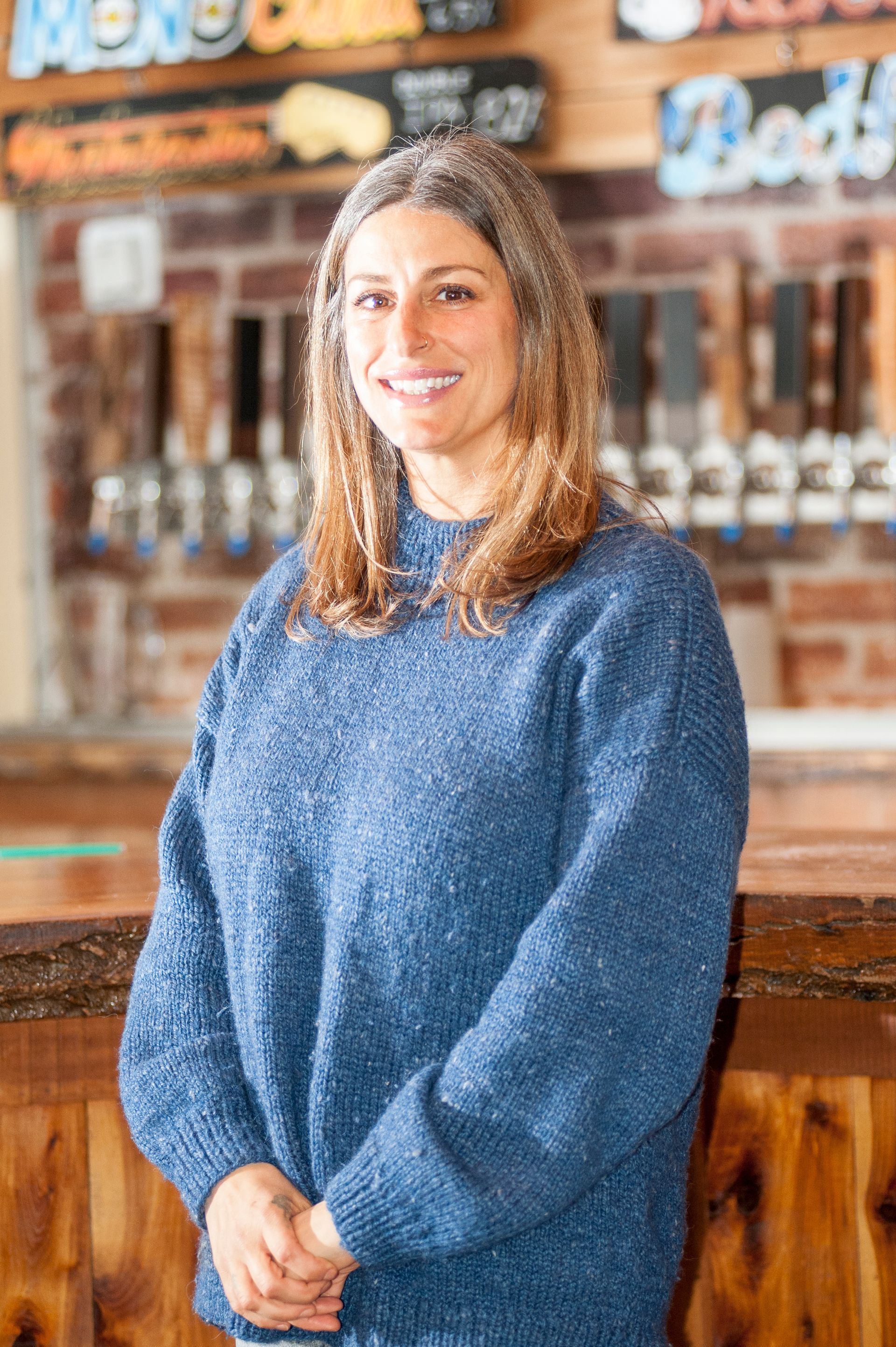 Woman smiles at the camera in a blue sweater, standing in front of a bar with beer taps.
