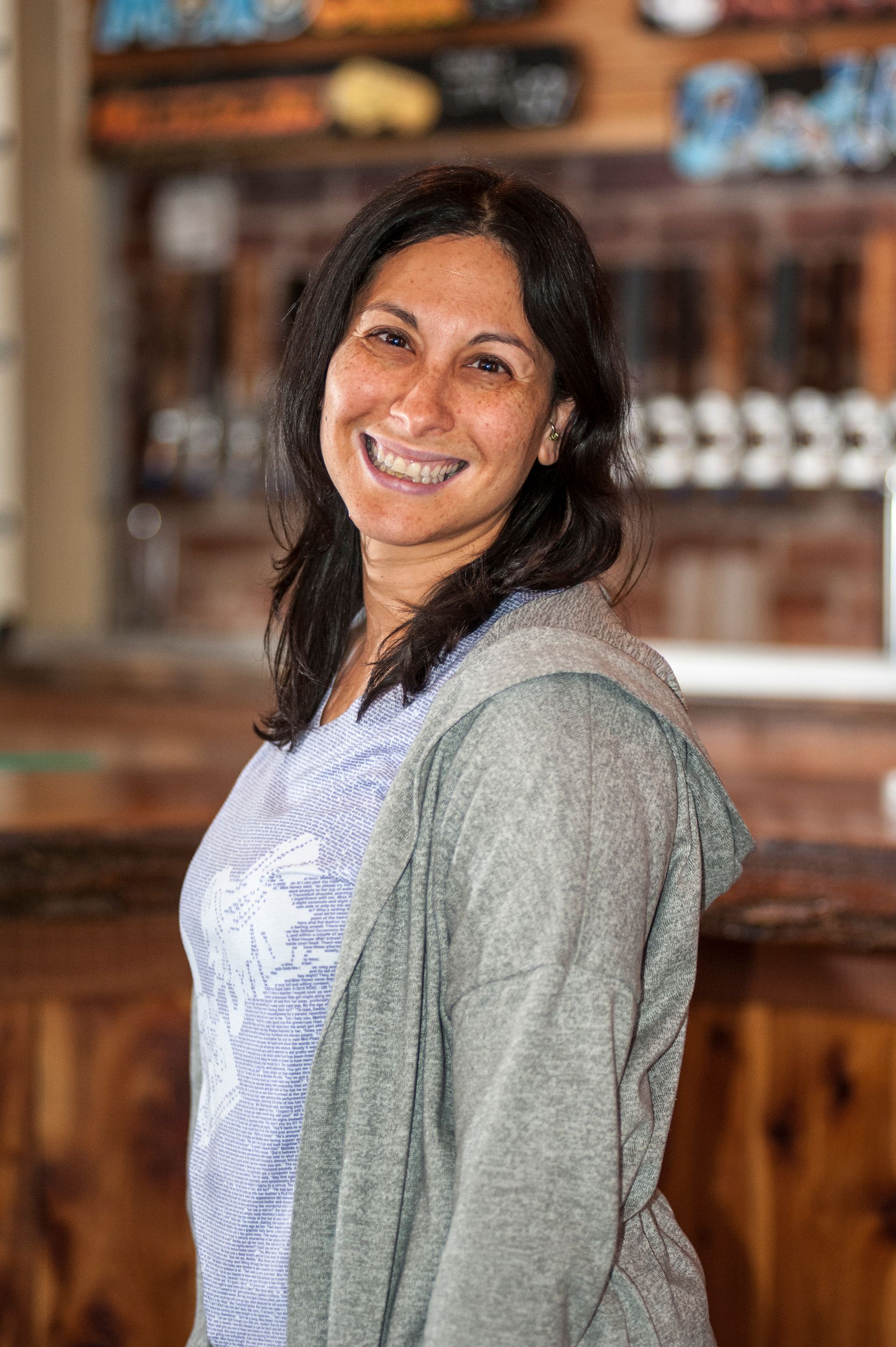 Woman smiling in a brewery, wearing a gray hoodie and blue patterned shirt, leaning towards the camera.