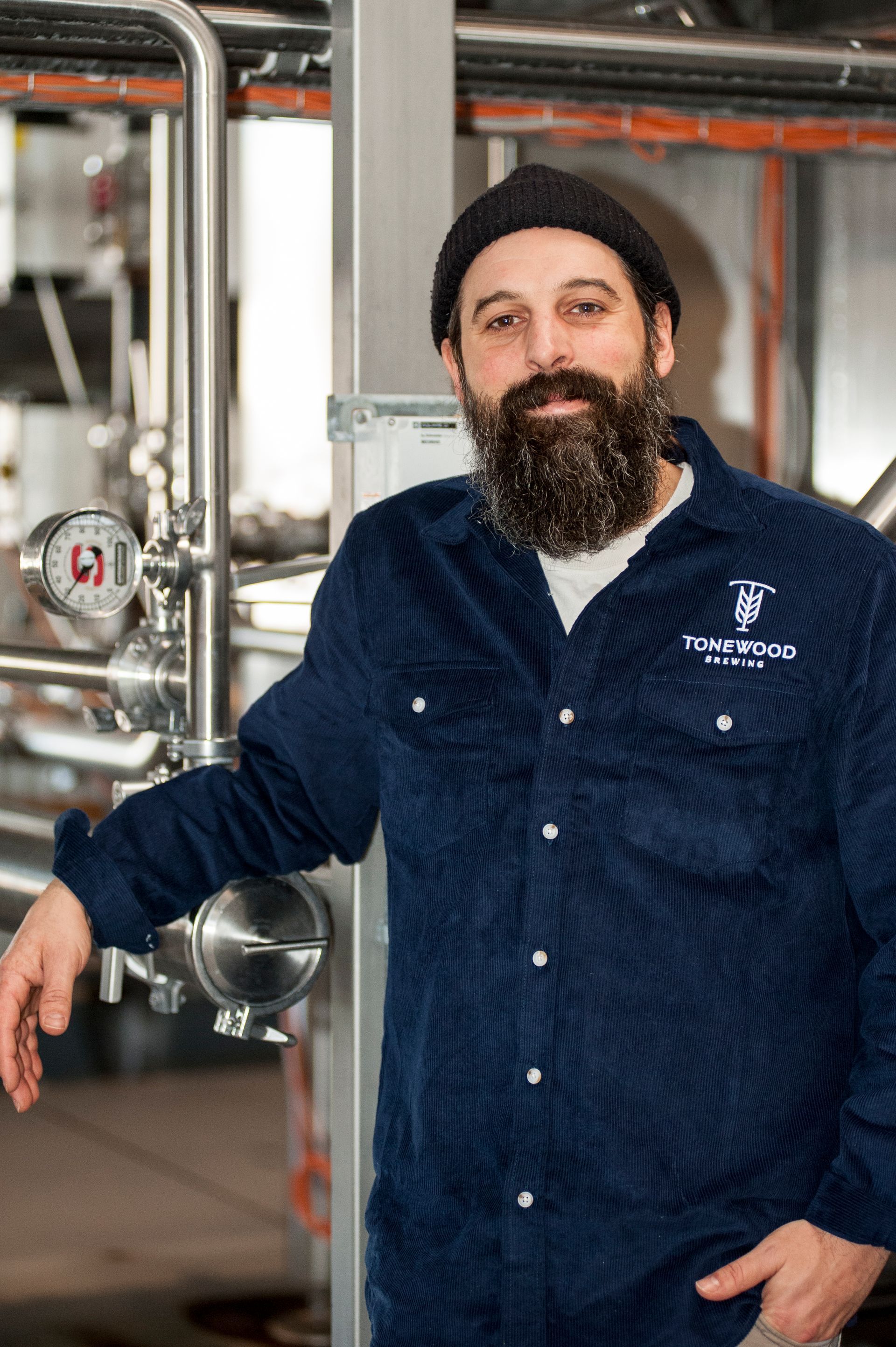 Man in blue shirt and beanie leans against brewery equipment.