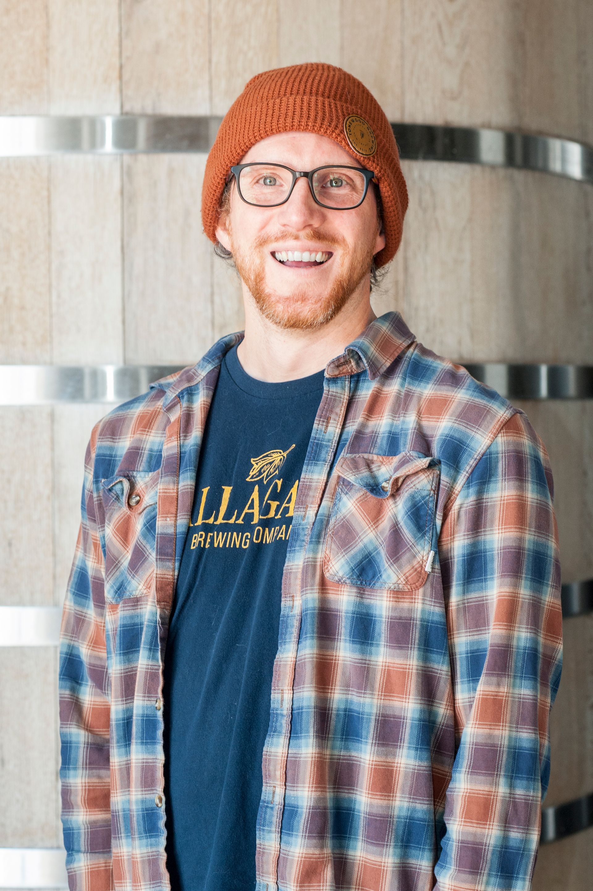 Man in orange hat and plaid shirt smiles in front of a wooden barrel.