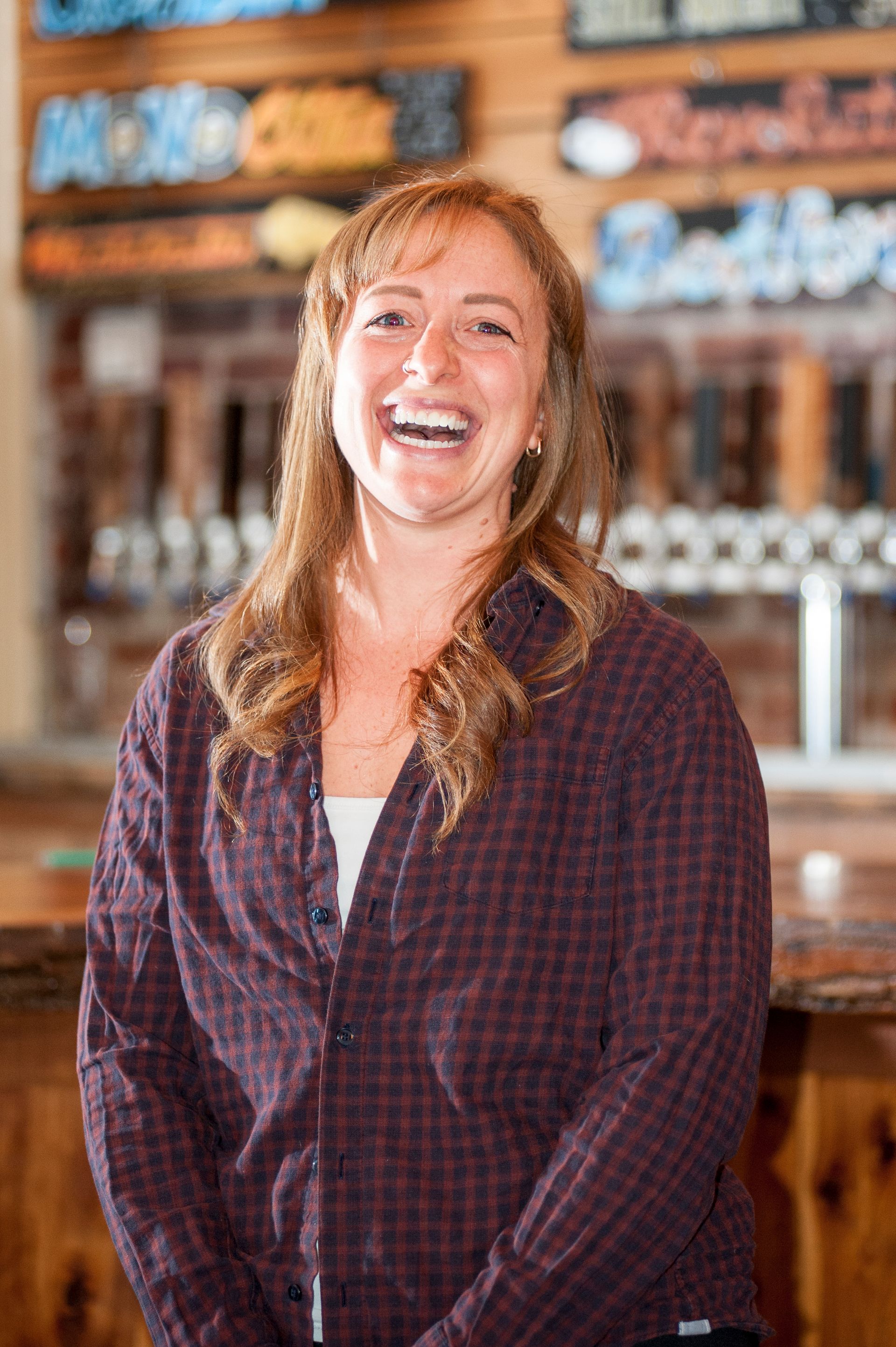 Woman laughing, wearing a checkered shirt, in a bar with beer taps in the background.
