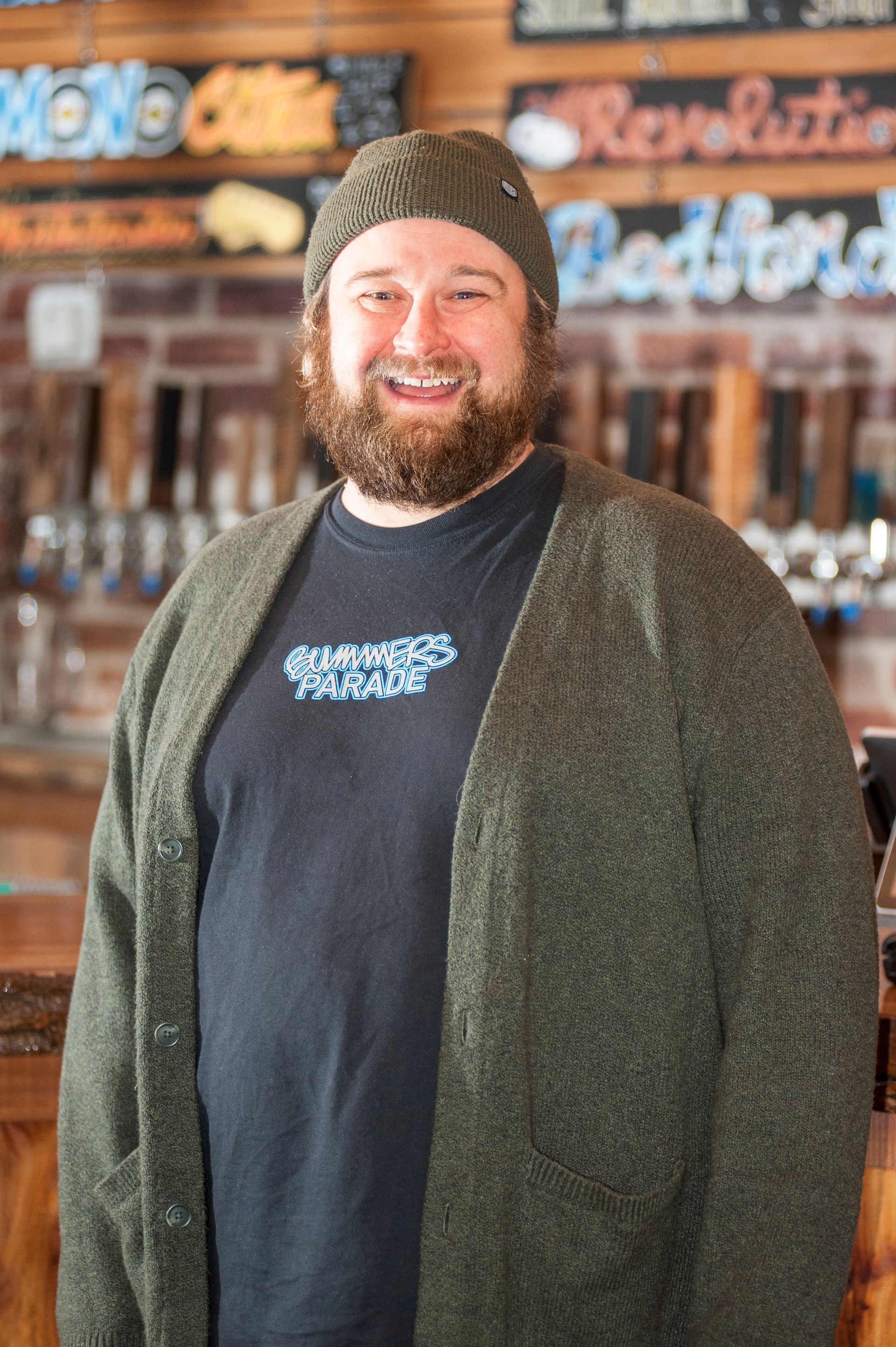 Man with beard and knit hat smiling, wearing a cardigan over a t-shirt, standing in a bar.