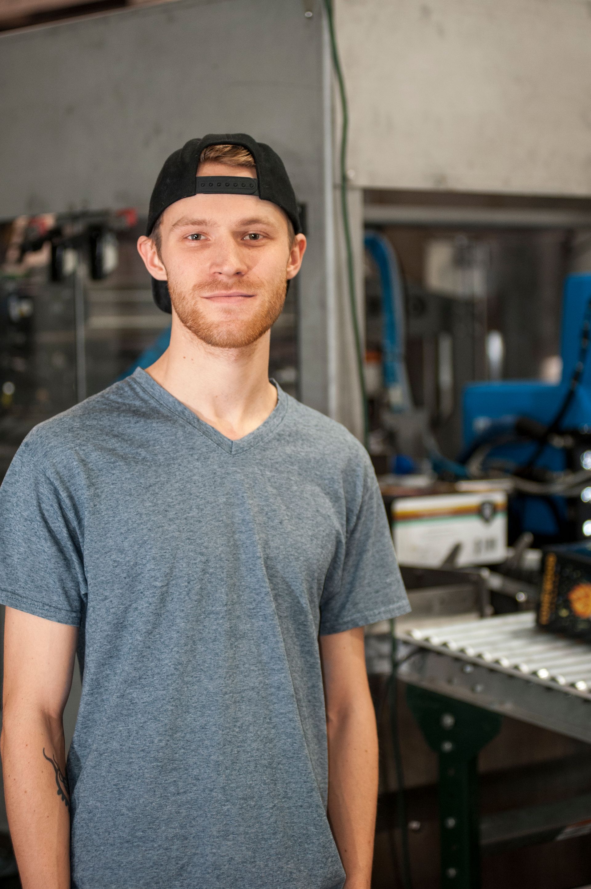 Man wearing a black baseball cap and gray V-neck shirt, smiles in a factory setting.
