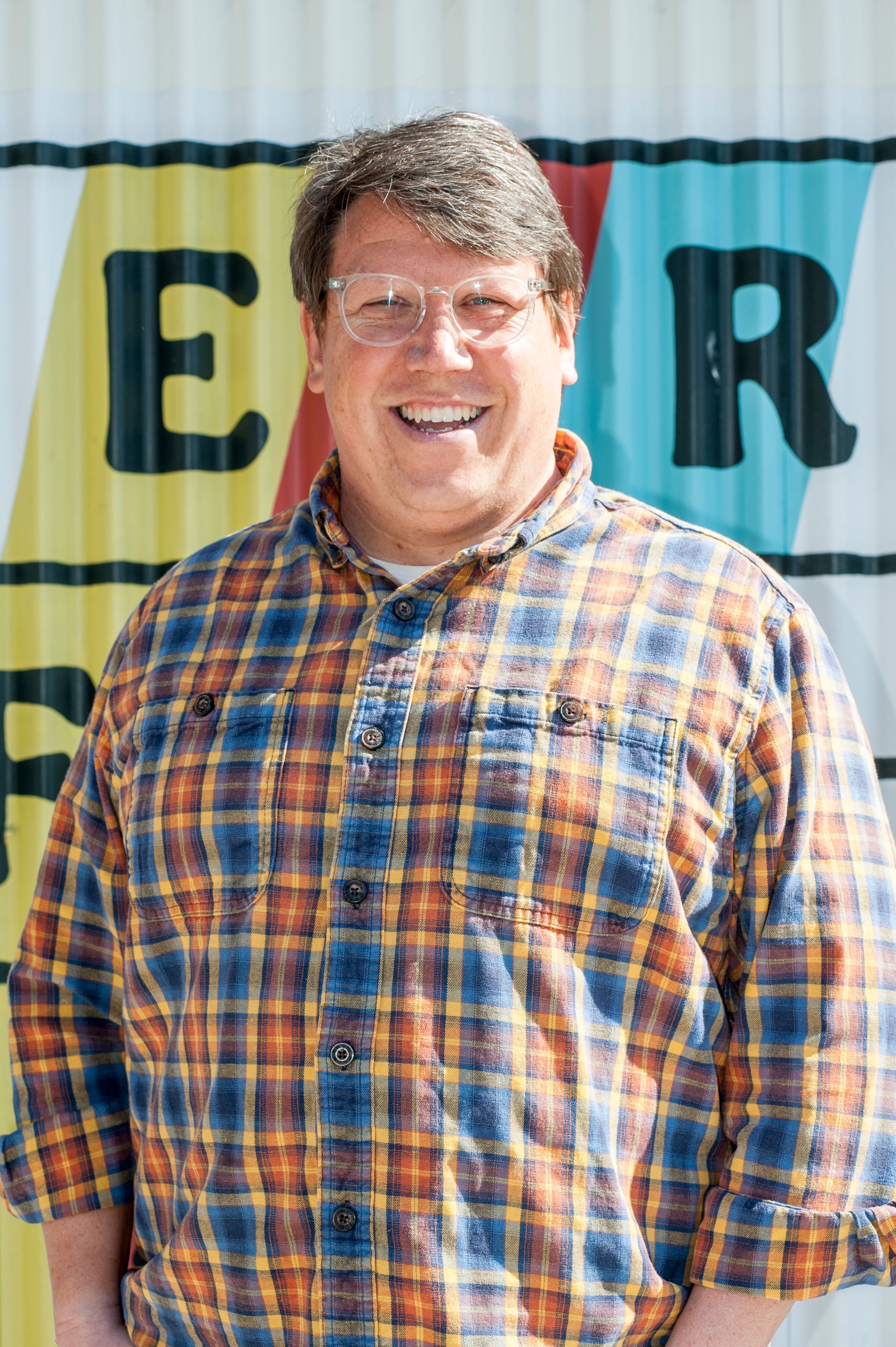 Man in plaid shirt smiling in front of colorful wall with lettering.