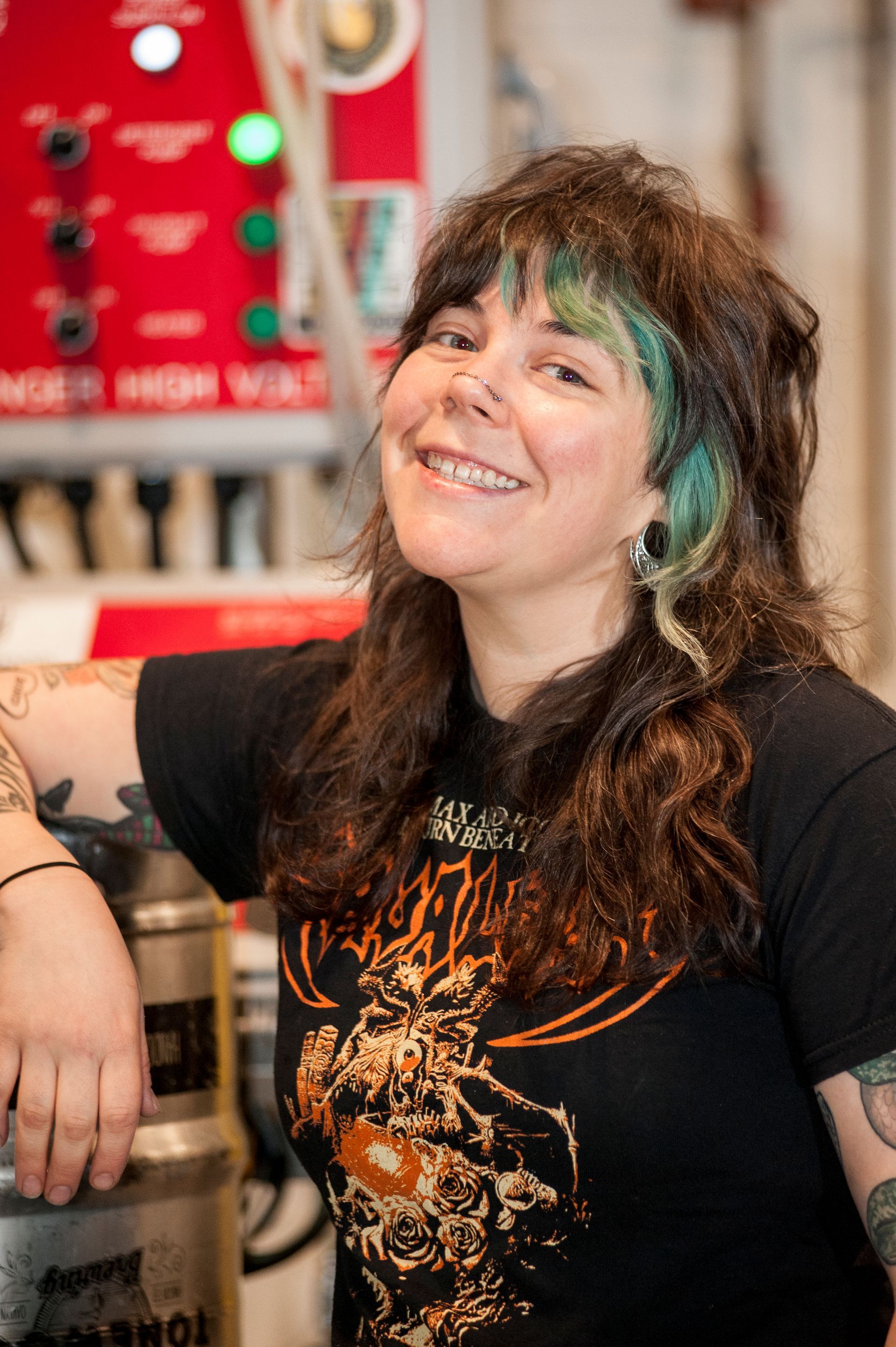 Woman with tattoos and green-streaked hair smiles, leaning on a metal tank. She wears a black graphic t-shirt in a industrial setting.