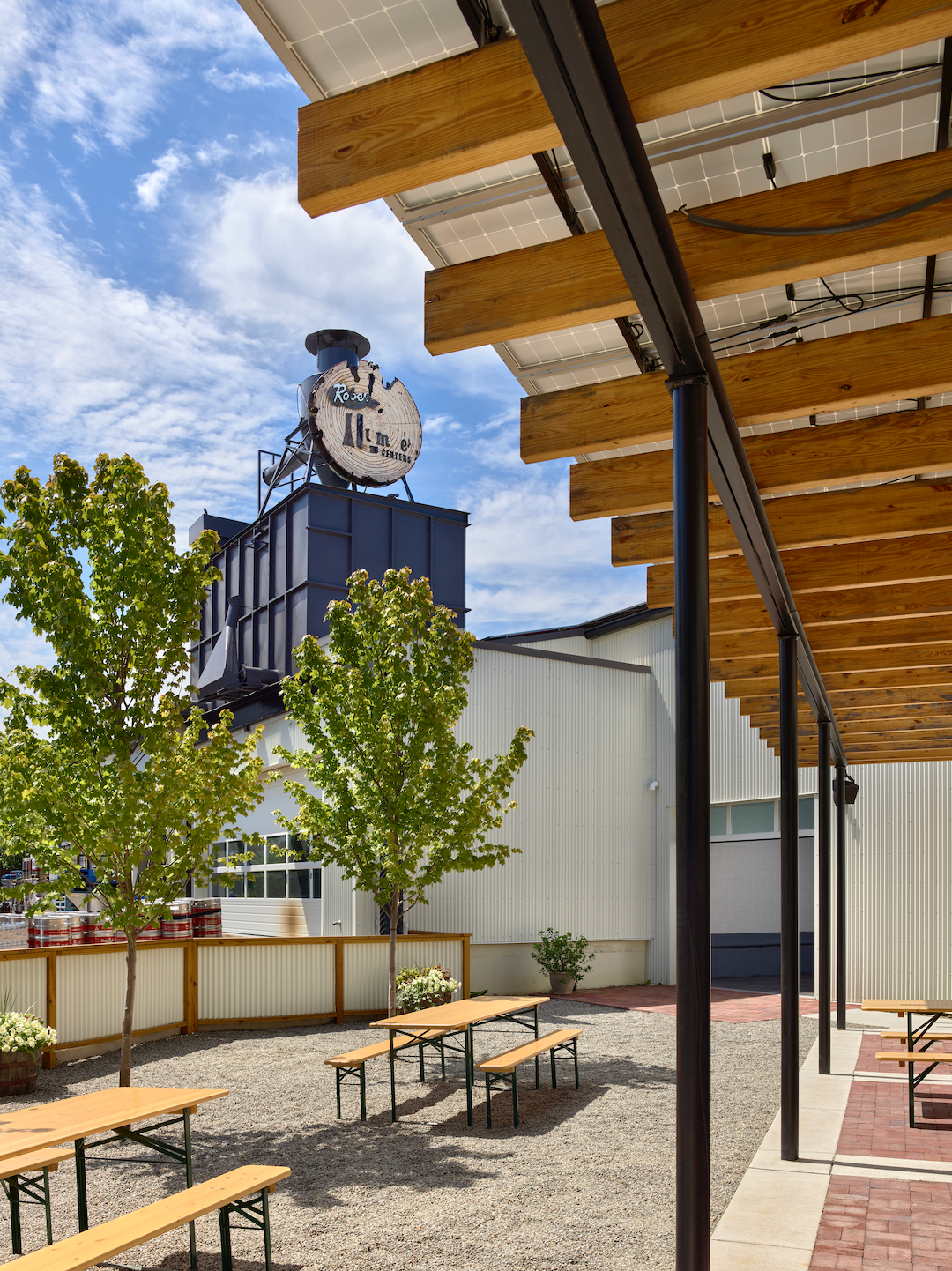 Outdoor patio with picnic tables under a solar panel roof, next to a building with a clock tower and trees.