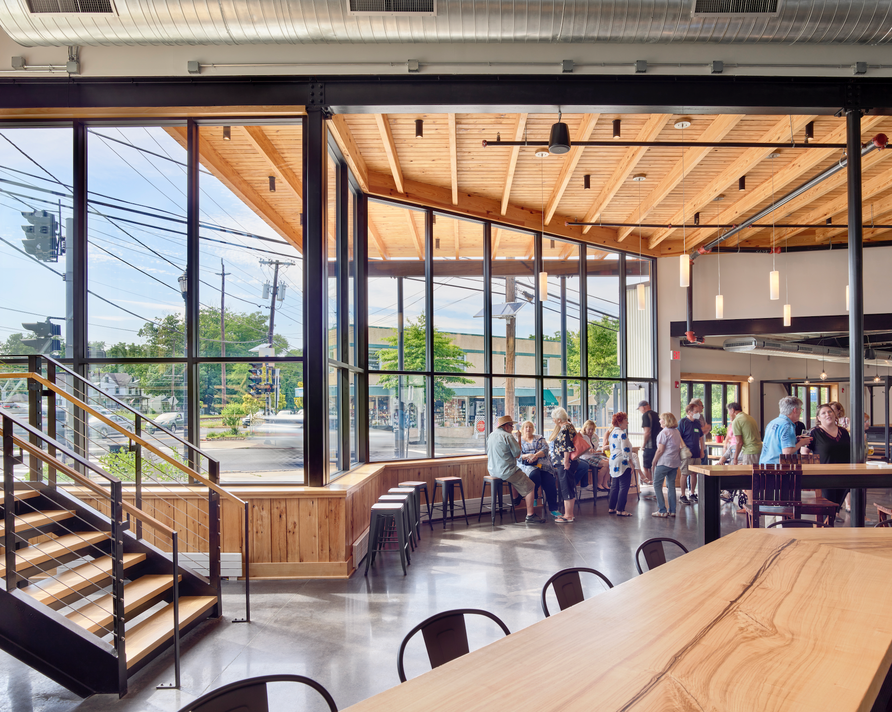 Interior of a modern building with large windows, wooden ceiling, and people gathered inside and outside.