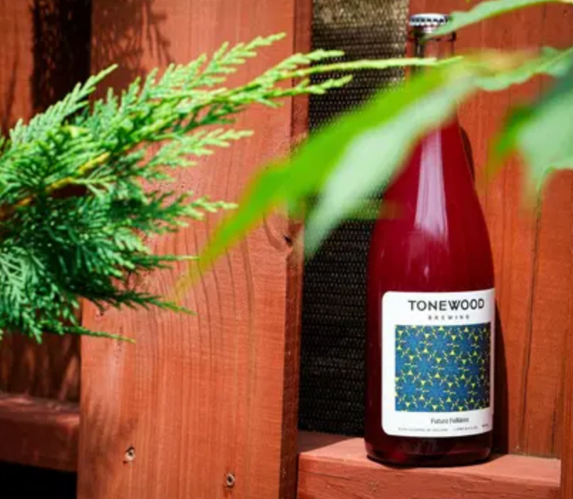 Bottle of red liquid with label, behind a cedar plant and wooden structure.