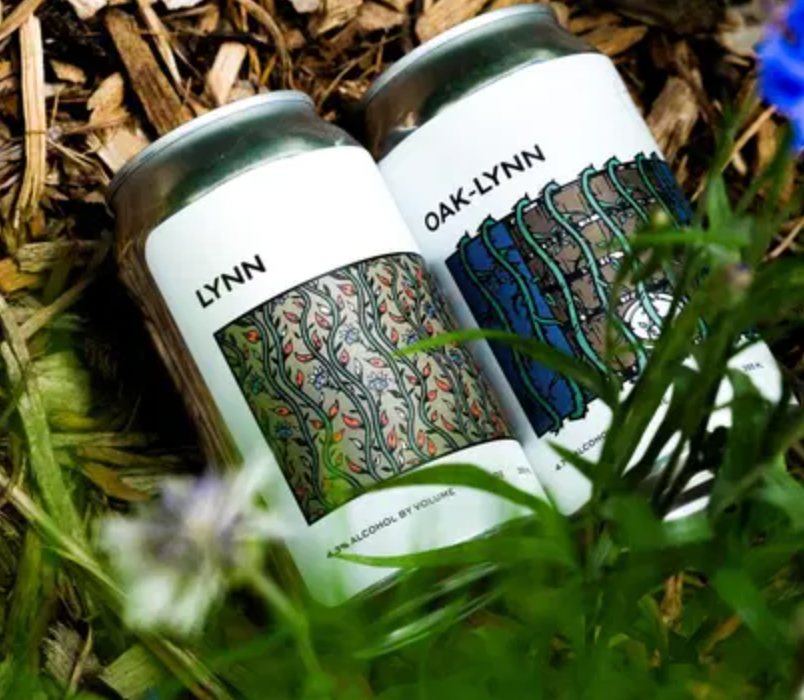 Two white beer cans with nature-themed labels, partially obscured by green plants and woodchips.
