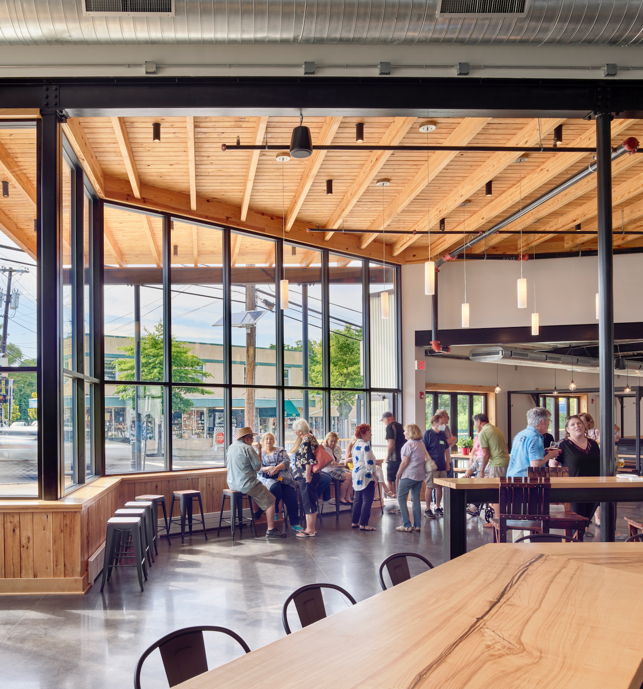 Interior of a modern building with large windows, people seated and standing at tables. Wooden ceiling and floor.