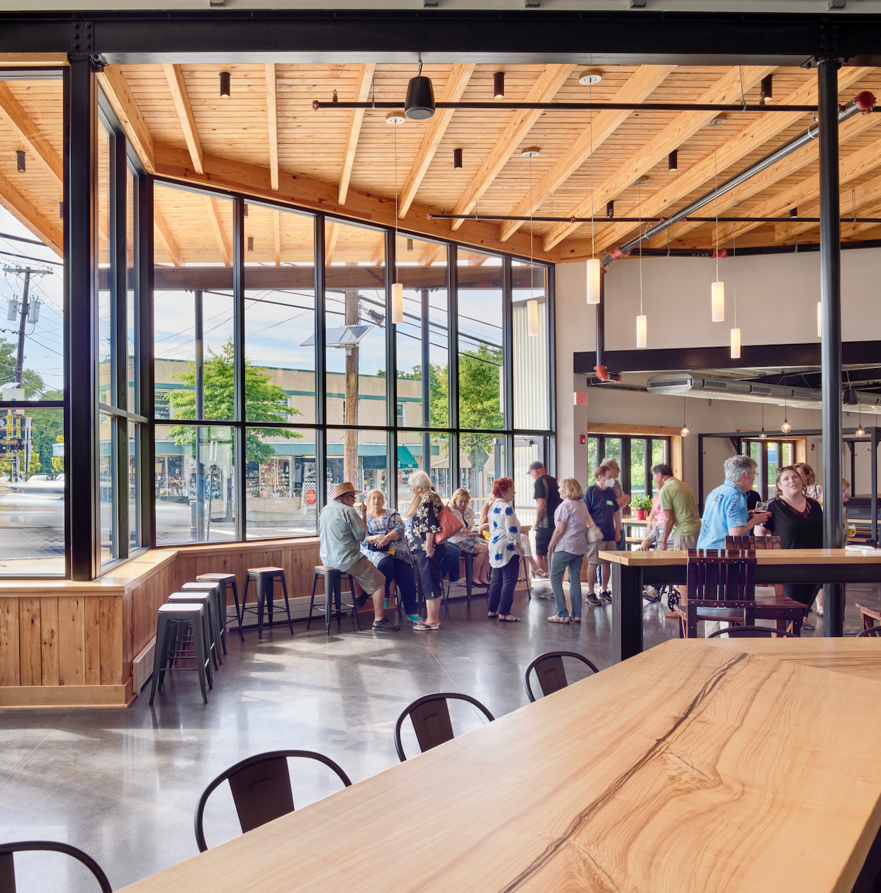 Interior of a modern building with large windows, people sitting at tables and bar. Wooden ceiling and floor.
