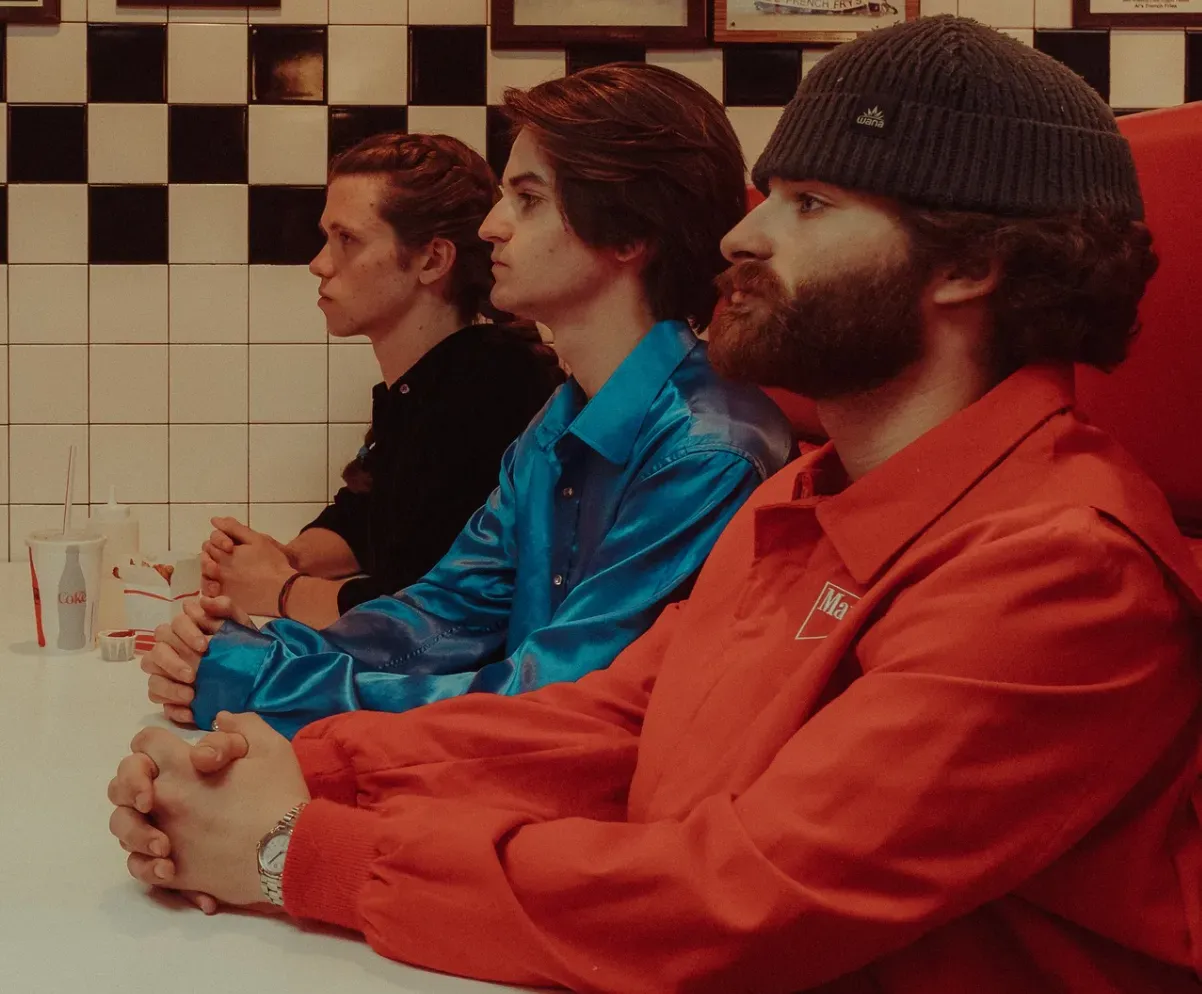 Three people sit at a diner counter. One wears a red shirt and beanie, the middle one a blue shirt, and the last a black shirt.