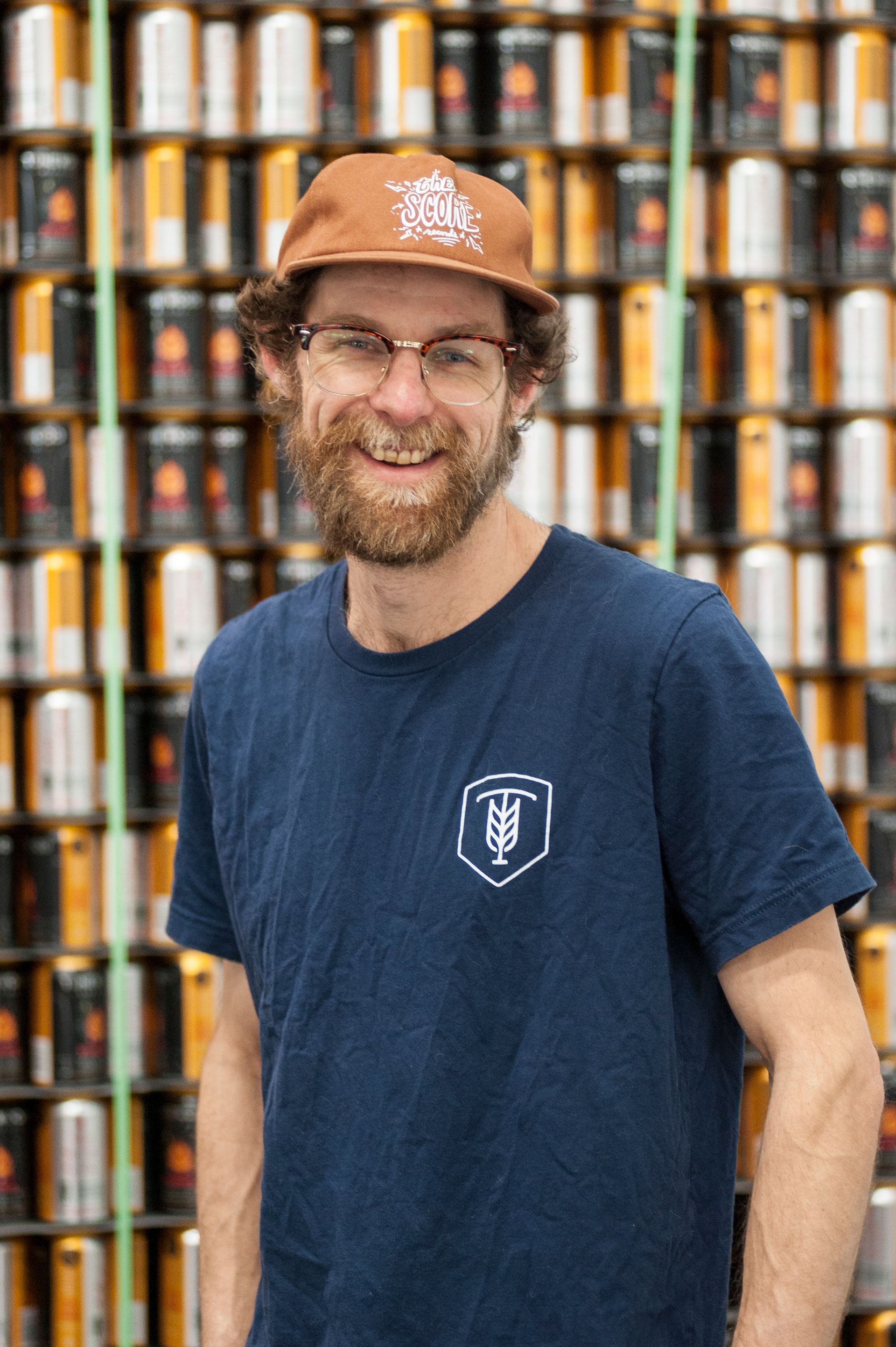 Man smiling in front of a wall of beer cans, wearing glasses and a hat.