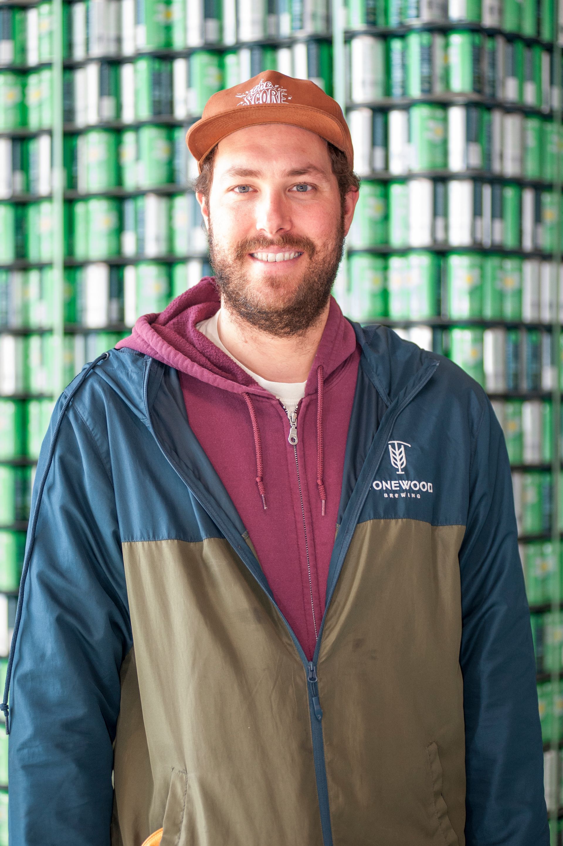 Man with beard and cap in front of a wall of green cans, wearing a blue, green, and red jacket.