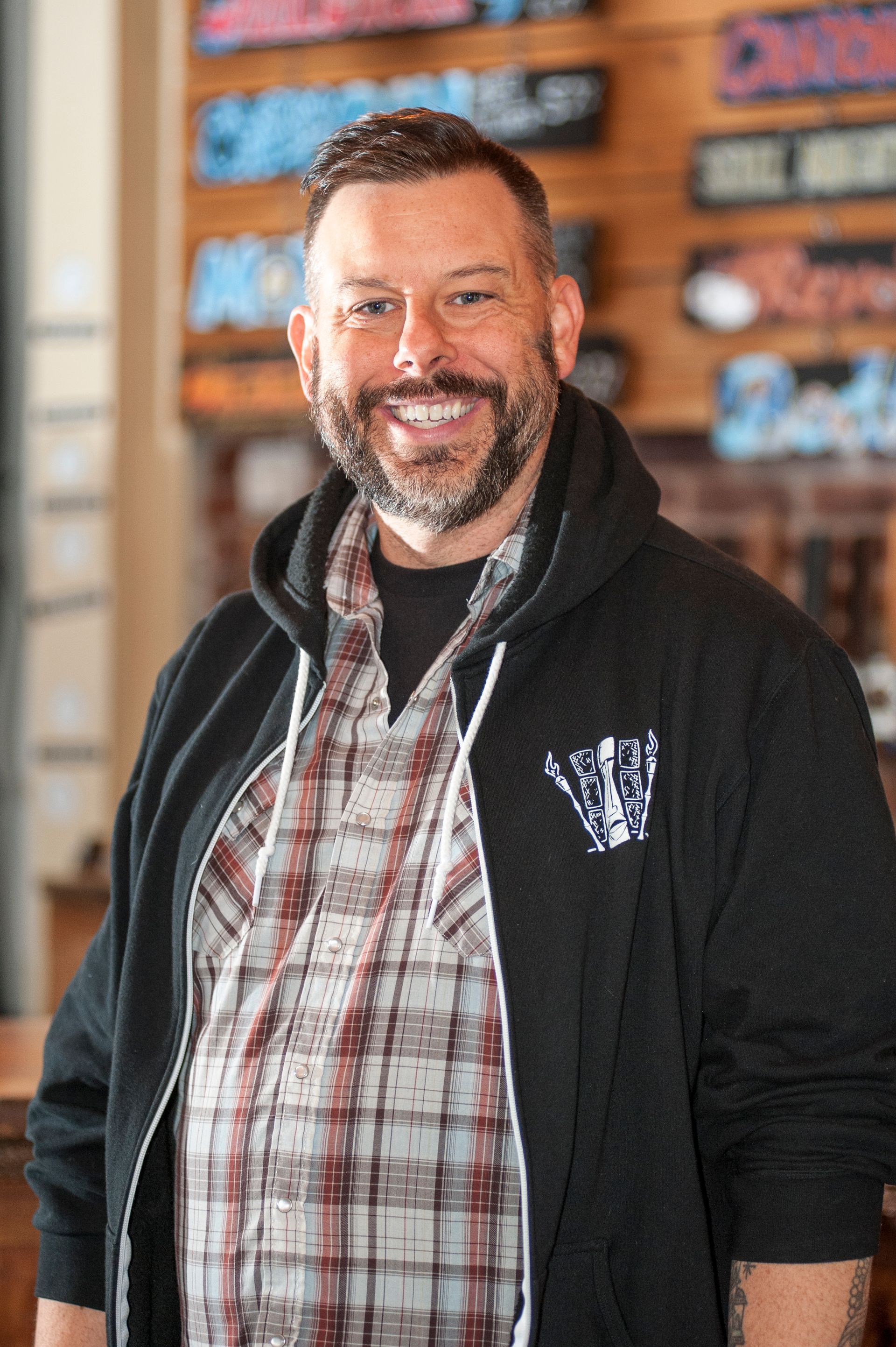 Man with a beard and a plaid shirt smiles, wearing a black hoodie in a bar setting.