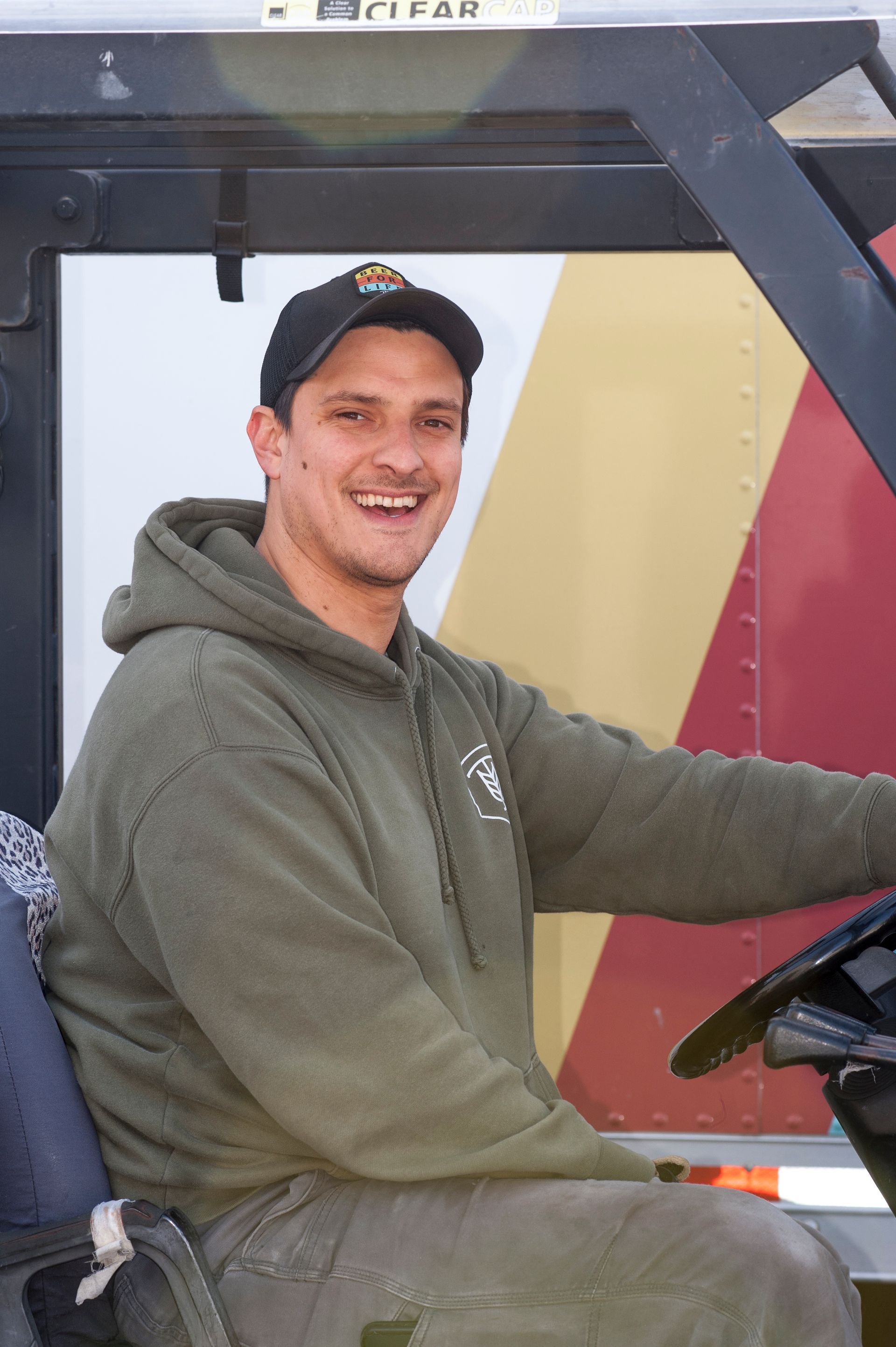 Man in green hoodie and cap, smiling while driving a vehicle with red and yellow accents.