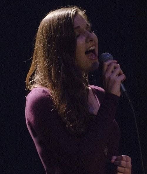 Woman singing into a microphone, mouth open, dark purple shirt, stage lighting.