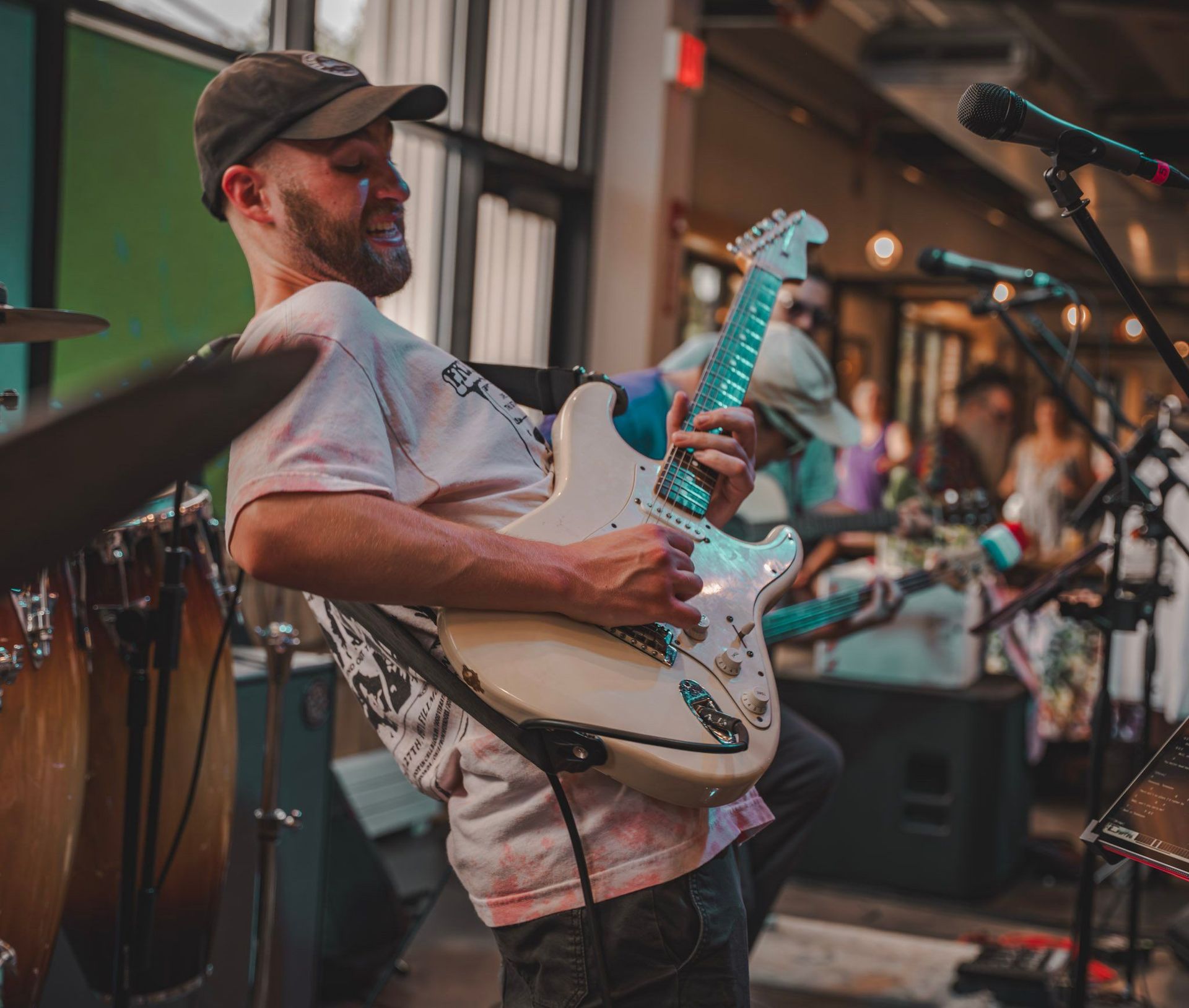 Man playing a white electric guitar on stage, wearing a cap and tie-dye shirt.