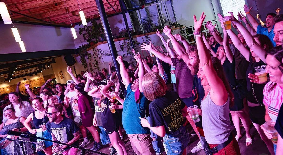Crowd with raised arms at a concert in a venue with stage lights.
