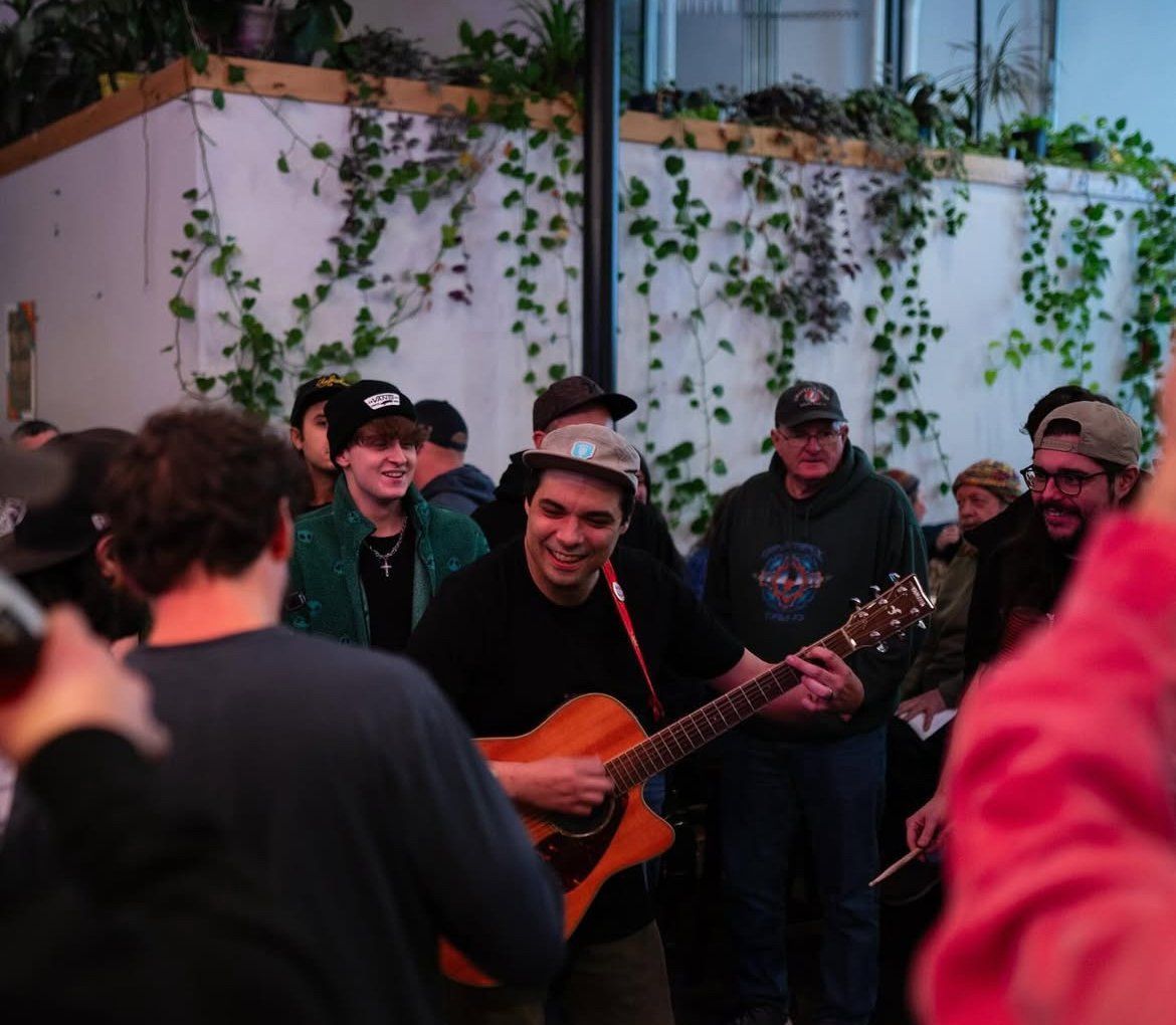 A musician plays acoustic guitar surrounded by smiling people in an indoor setting with plants.