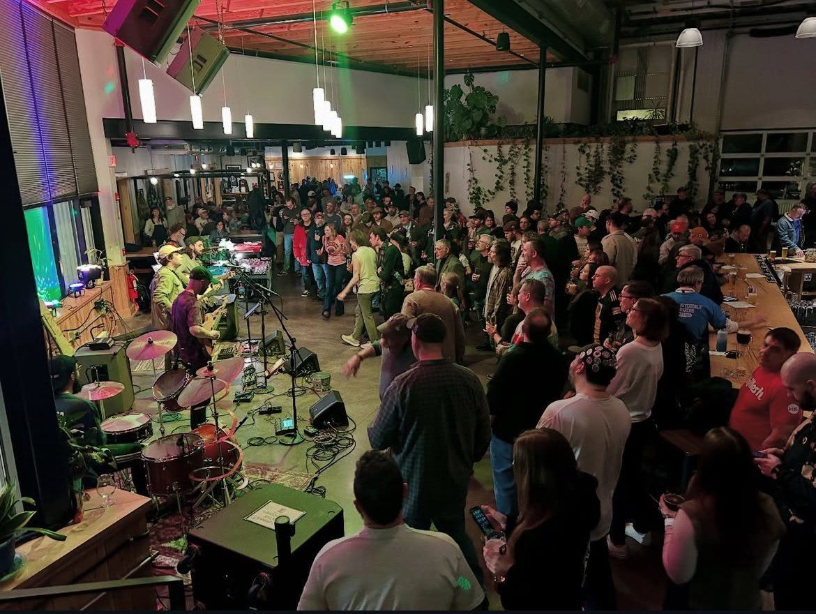Band performing on stage in a crowded venue. People stand, listen, and enjoy the show in a building with indoor plants.