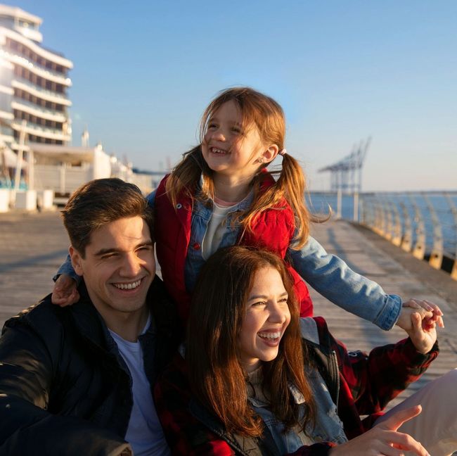 A man and woman are sitting on a pier with a little girl on their shoulders.