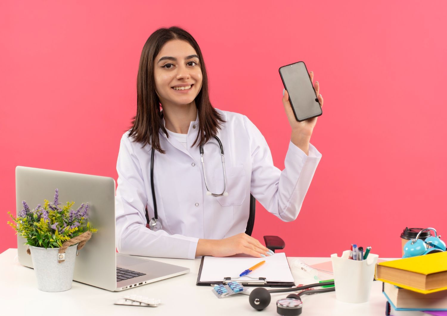 A female doctor is sitting at a desk holding a cell phone.