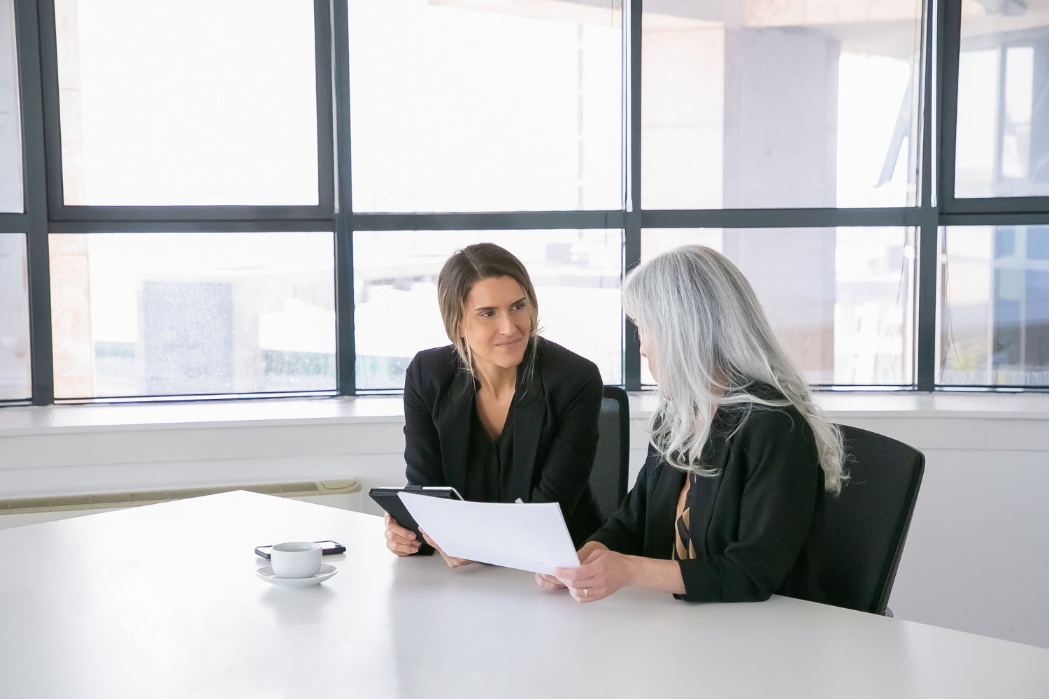 Two women in a bright office review documents at a table, smiling, one with gray hair.