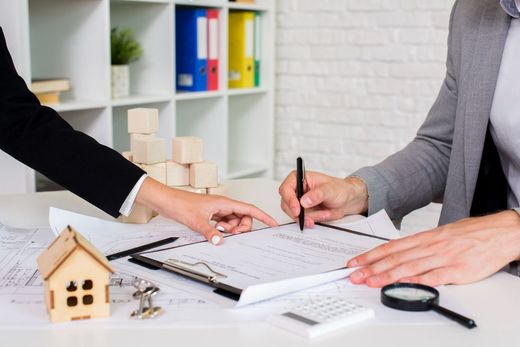 Person signing document at desk, woman pointing. Wooden house, keys, and calculator on the table.